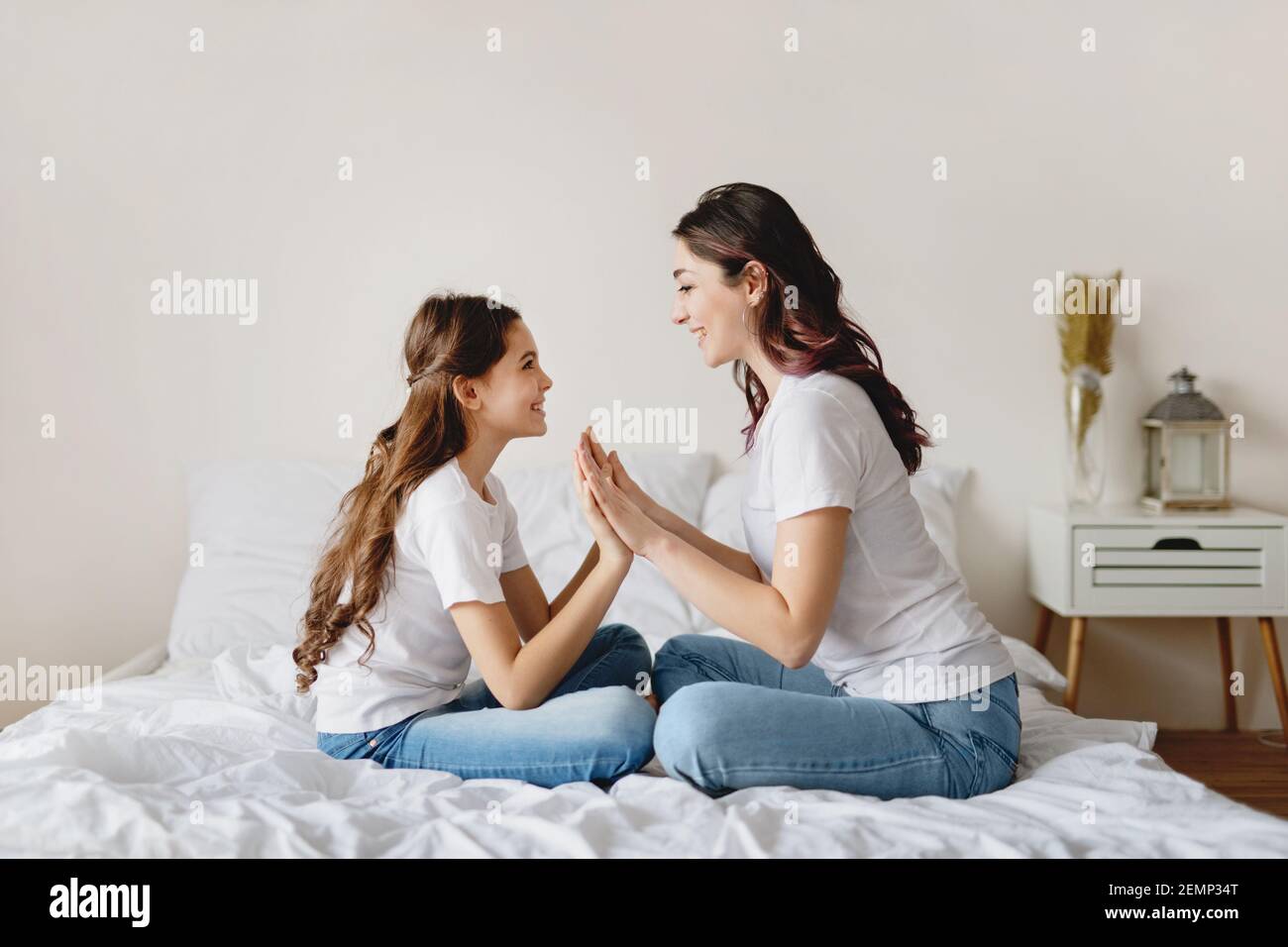 Nettes Mädchen und ihre schöne Mutter sitzen auf dem Bett und klatschen die Hände zusammen. Stockfoto