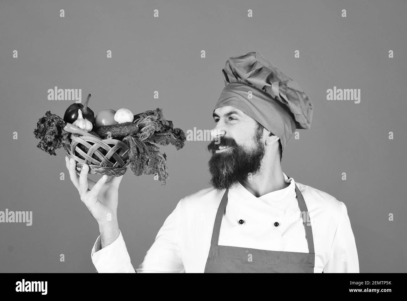 Mann mit Bart auf blauem Hintergrund. Kochen mit lächelndem Gesicht in burgunderrot Uniform hält Gemüse in Weidenschale. Der Küchenchef hält Salat, Tomaten, Pfeffer und Pilze bereit. Grünes Lebensmittelkonzept. Stockfoto