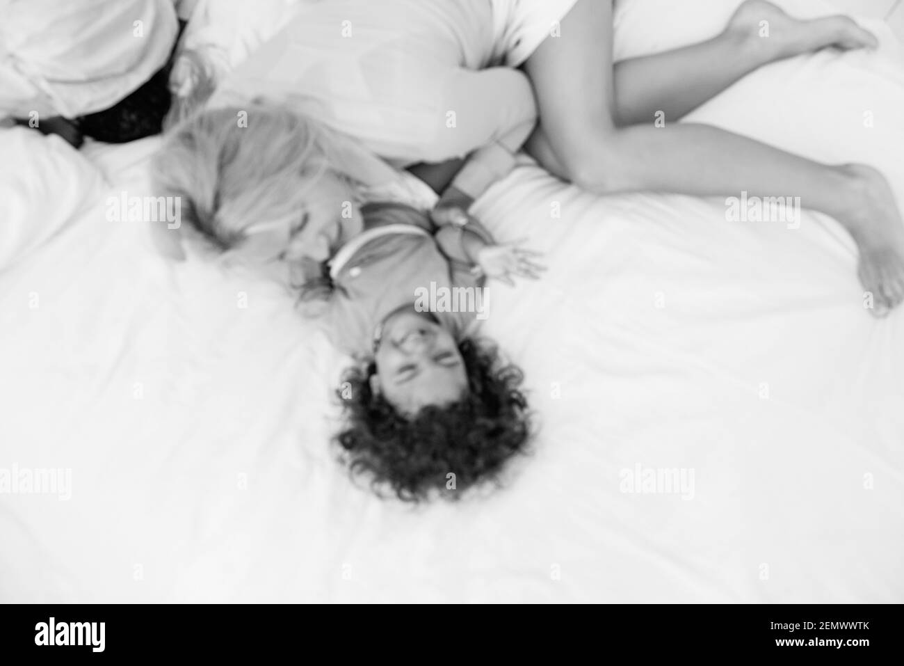 Defokusste junge kaukasische blonde Mutter kitzelt ihre kleine afroamerikanische Tochter. Auf weißem Bett liegen, Spaß haben, Blick von oben. Die Mädchen lachen, umarmen, die Stockfoto
