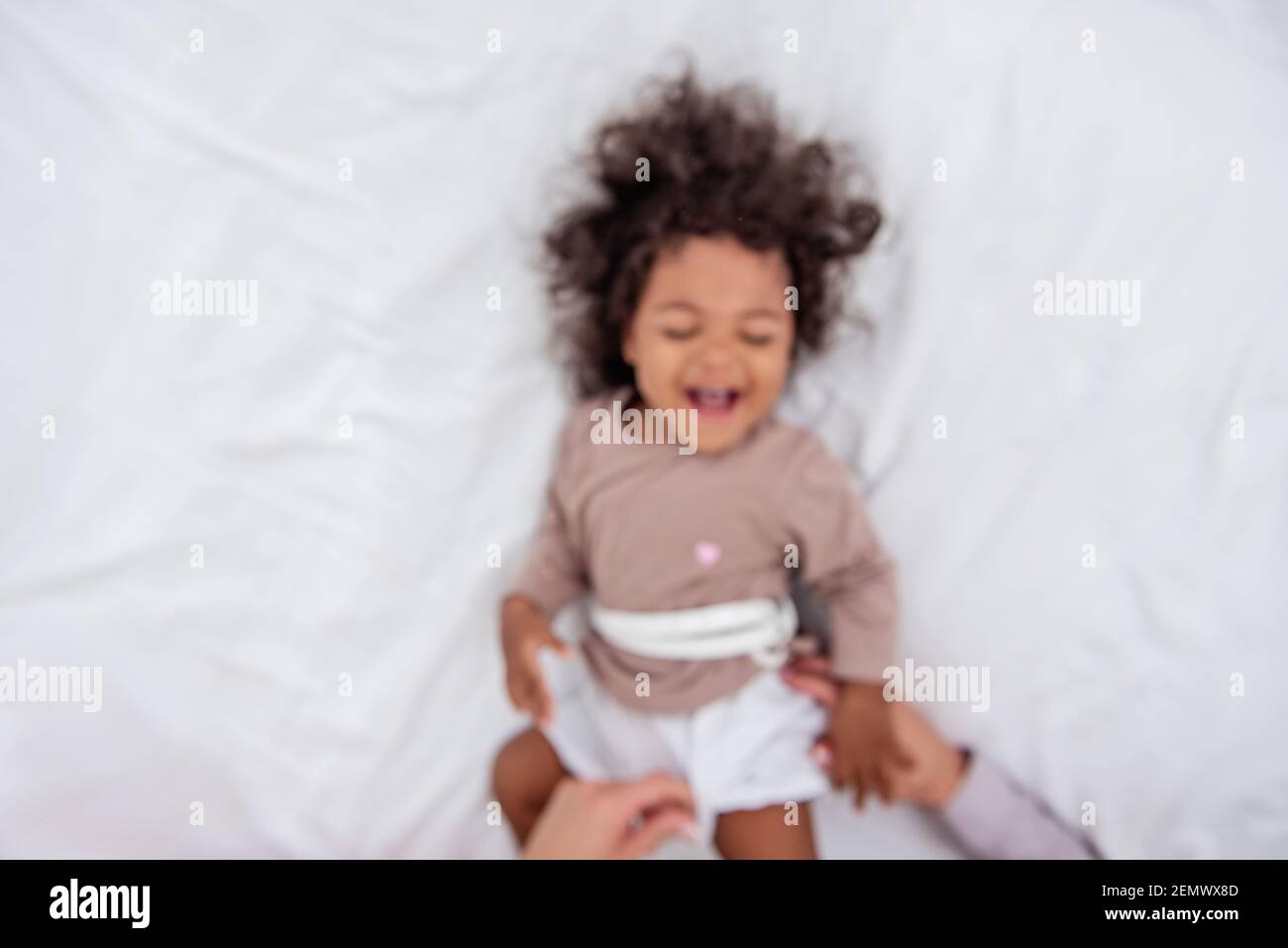 Defokusste junge kaukasische blonde Mutter kitzelt ihre kleine afroamerikanische Tochter. Auf weißem Bett liegen, Spaß haben, Blick von oben. Die Mädchen lachen, umarmen, die Stockfoto