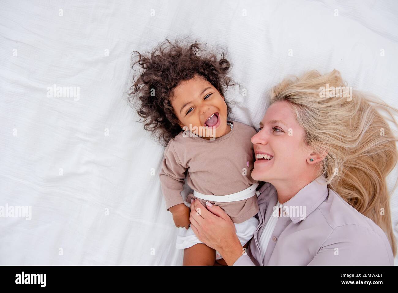 Junge kaukasische blonde Mutter kitzelt ihre kleine afroamerikanische Tochter. Auf weißem Bett liegen, Spaß haben, Blick von oben. Die Mädchen lachen, umarmen, die Frau kis Stockfoto