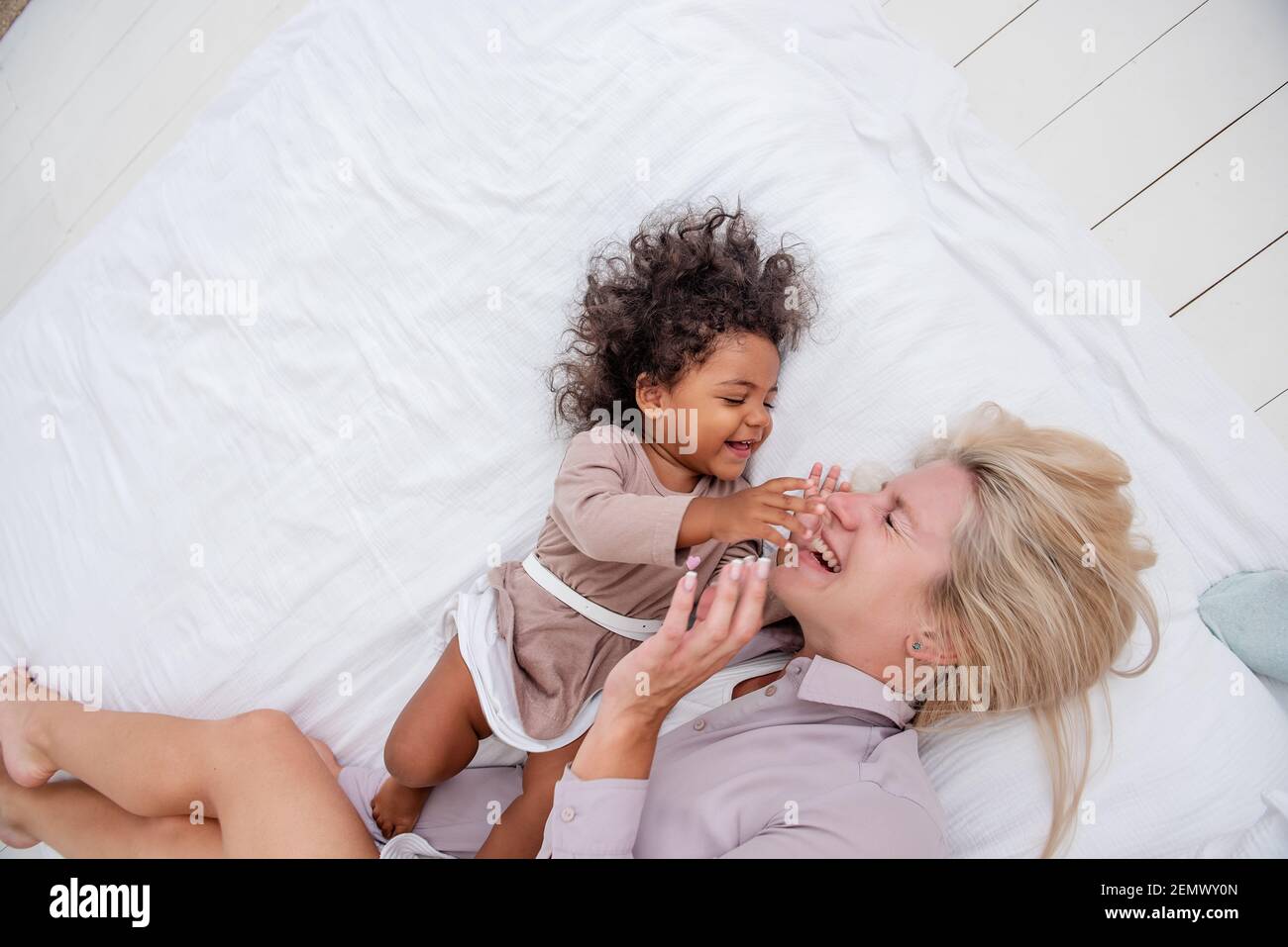 Junge kaukasische blonde Mutter kitzelt ihre kleine afroamerikanische Tochter. Auf weißem Bett liegen, Spaß haben, Blick von oben. Die Mädchen lachen, umarmen, die Frau kis Stockfoto
