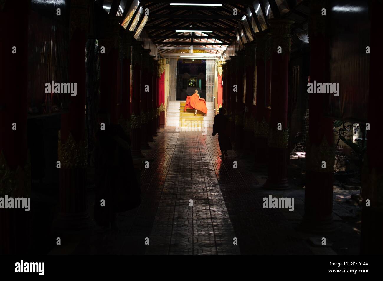 YANGON, MYANMAR - JANUAR 1 2020: Ein junger Mönch, der die traditionelle orangefarbene Robe fixiert, während er im Sonnenschein einen langen dunklen Gehweg hinuntergeht Stockfoto