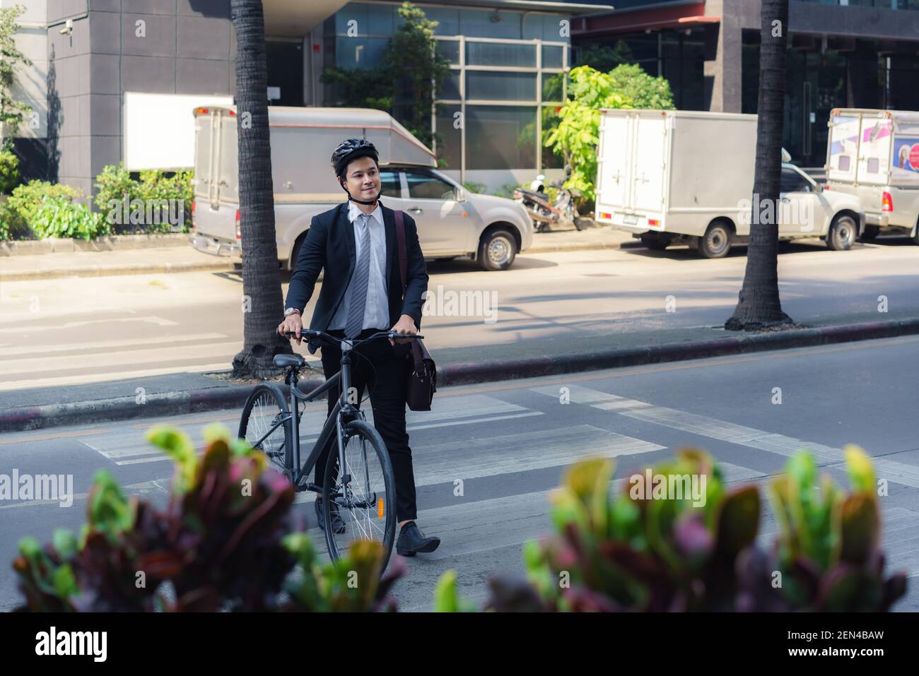 Ein asiatischer Geschäftsmann schiebt ein Fahrrad über einen Fußgängerüberweg auf einer Stadtstraße während eines morgendlichen Pendelwegs. Stockfoto