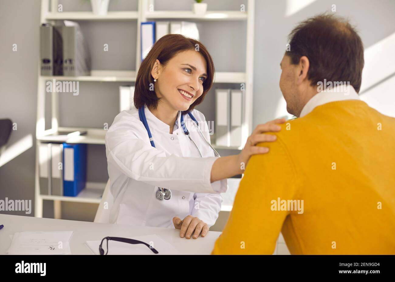 Lächelnder Arzt berührt die Schulter des älteren Mannes, unterstützt ihn und begrüßt ihn Er in der Klinik Stockfoto