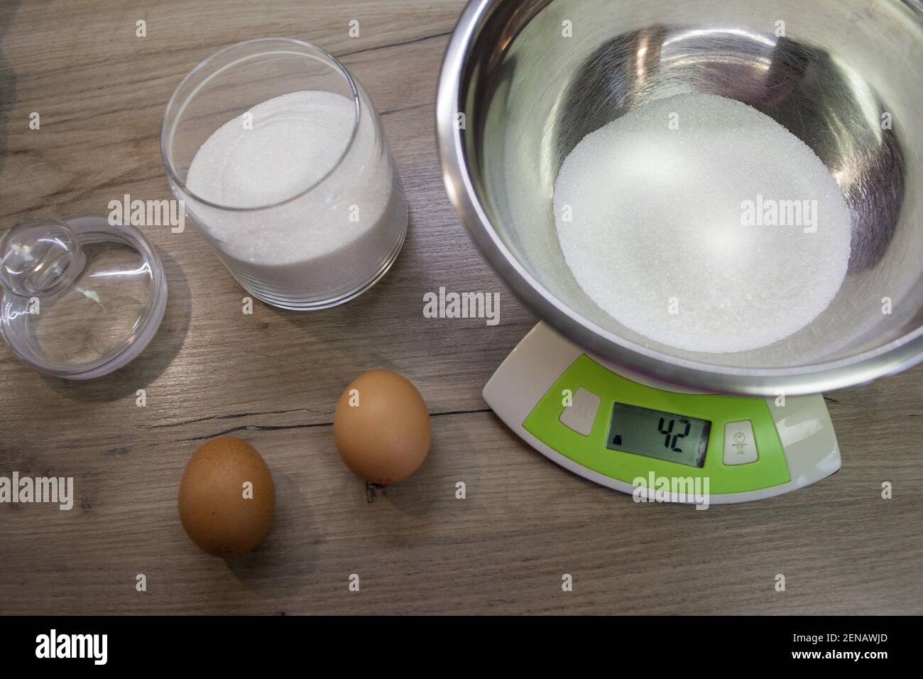 Weißer Zucker in einer Schüssel auf der Waage und zwei Eier auf dem Holzhintergrund. Der zweite Schritt für die Herstellung von Muffins, Kuchen, Keks. Backvorgang Schritt für Schritt Stockfoto