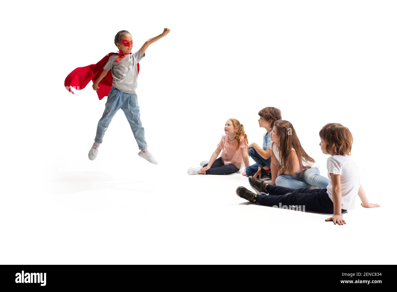 Leistung. Kind, das vorgibt, ein Superheld zu sein, mit ihren Freunden, die um sie herum sitzen. Kinder aufgeregt, inspiriert von ihrem starken, tapferen Freund in rotem Mantel isoliert auf weißem Hintergrund. Träume, Emotionen Konzept. Stockfoto