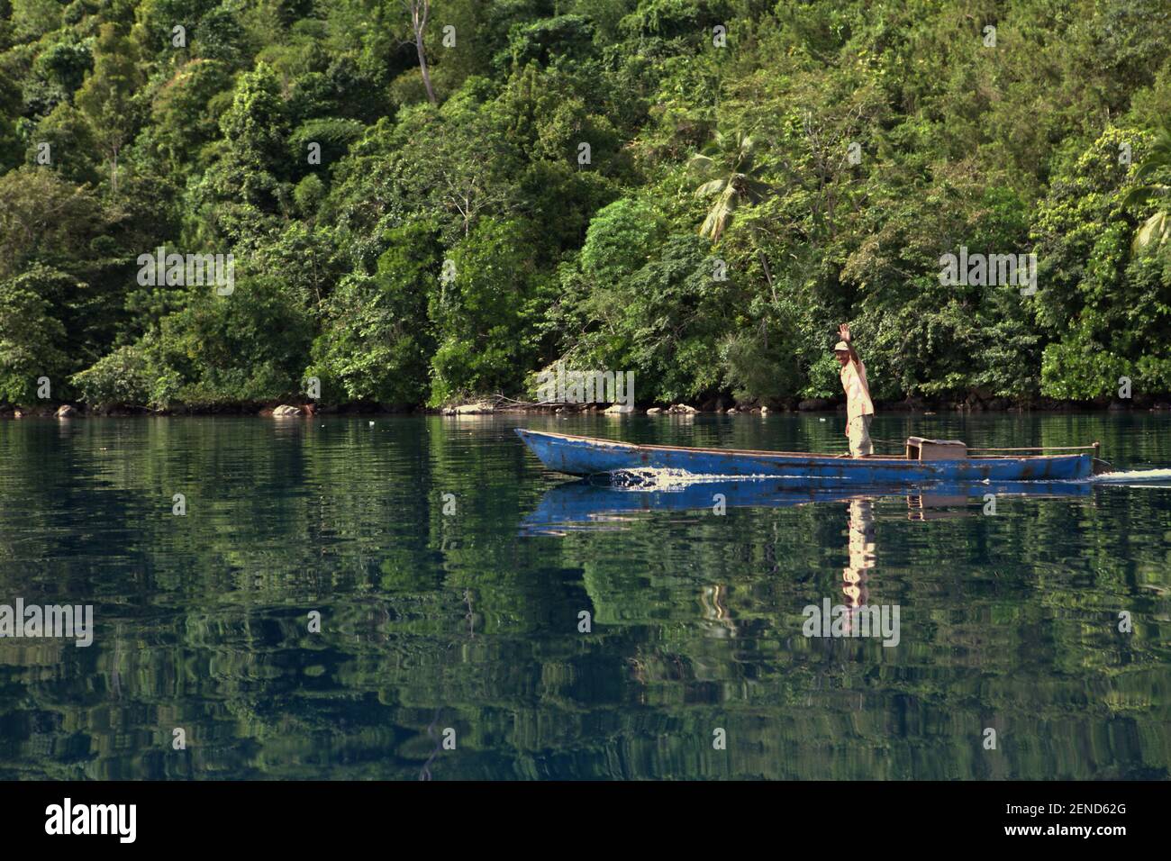 Ein Mann begrüßt vom Holzboot aus, während er auf dem Küstenwasser in der Nähe einer Kalksteinklippe in der Nähe des Öko-Tourismus-Dorfes Sawai in Central Maluku, Maluku, Indonesien segelt. Stockfoto