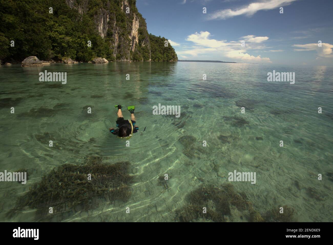 Ein Schnorchler schwimmt auf dem Küstenwasser und achtet auf den Meeresboden, der teilweise mit Korallenriffen bedeckt ist, fotografiert im Hintergrund eines Kalksteinhügels in der Nähe des Dorfes Sawai im Zentrum von Maluku, Provinz Maluku, Indonesien. Stockfoto