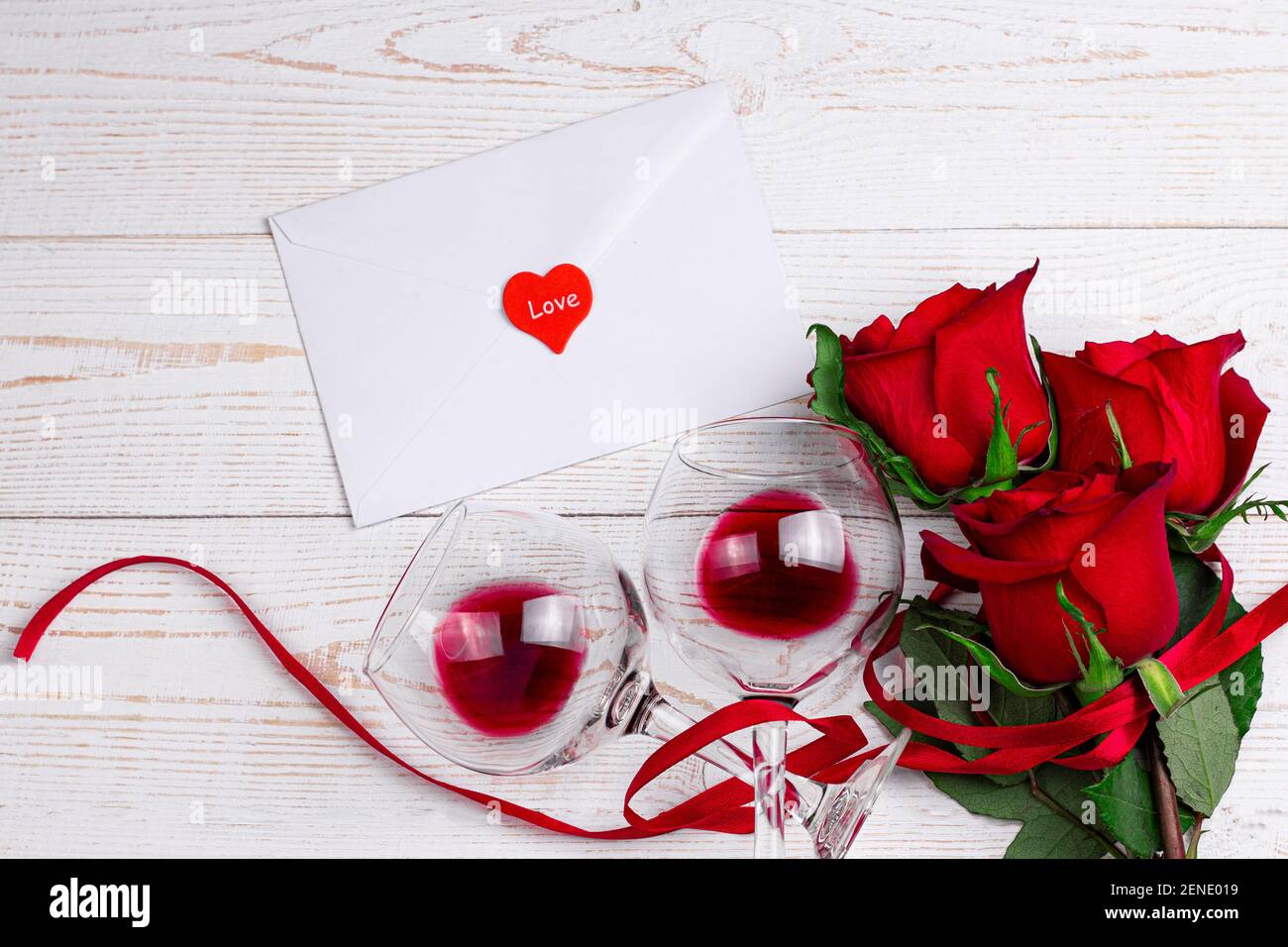 Weingläser mit Rotwein, Umschlag und roten Rosen mit rotem Band auf weißem Holzhintergrund. Valentinstag romantisches Abendessen flat Lay Top View Konzept Stockfoto