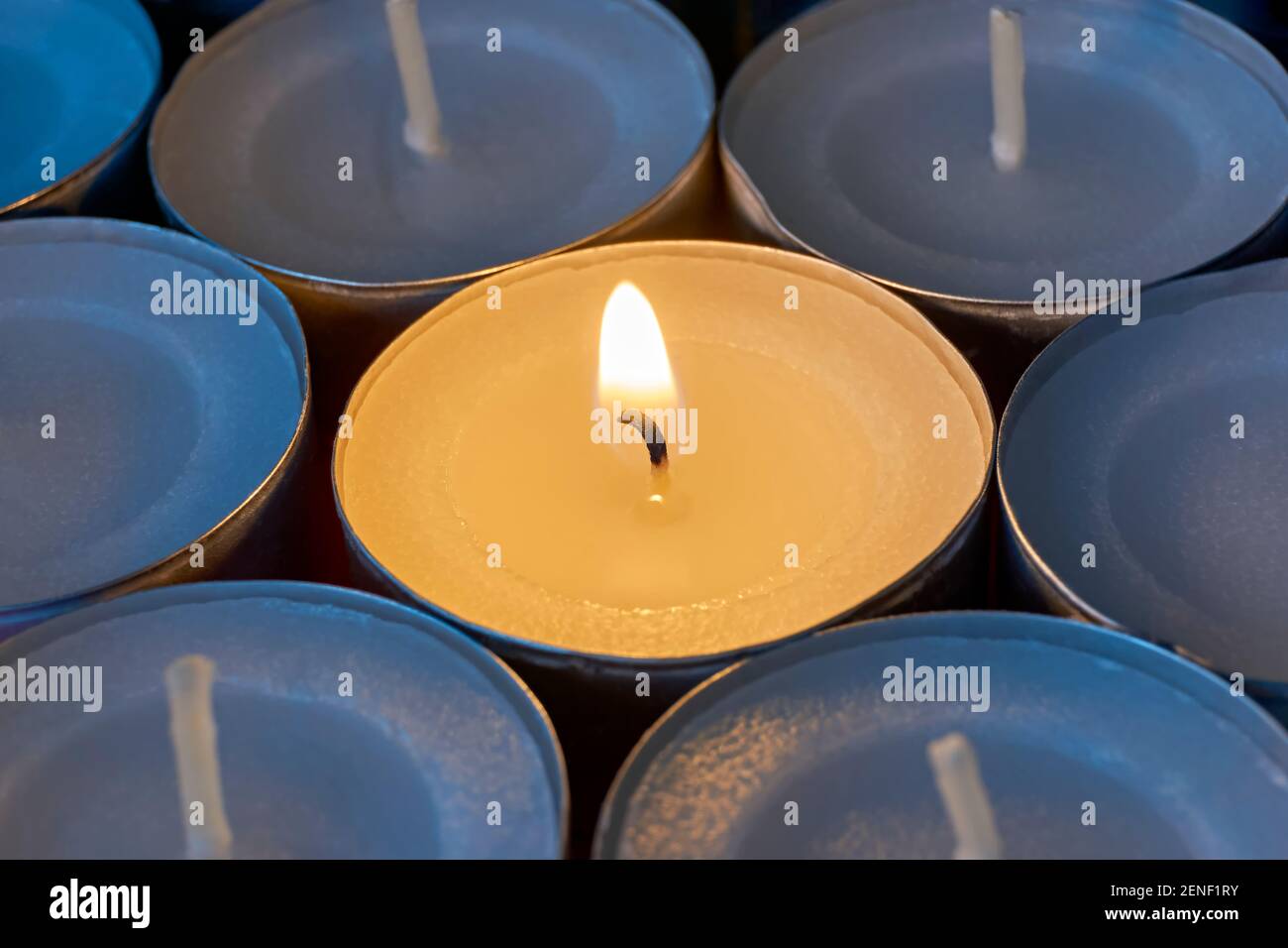Nahaufnahme von Teelampen mit einer beleuchtet. Eine einzelne warme orange Flamme zwischen Kerzen im blauen Schatten. Weihnachten, Beileid oder Begräbnishintergrund. Stockfoto