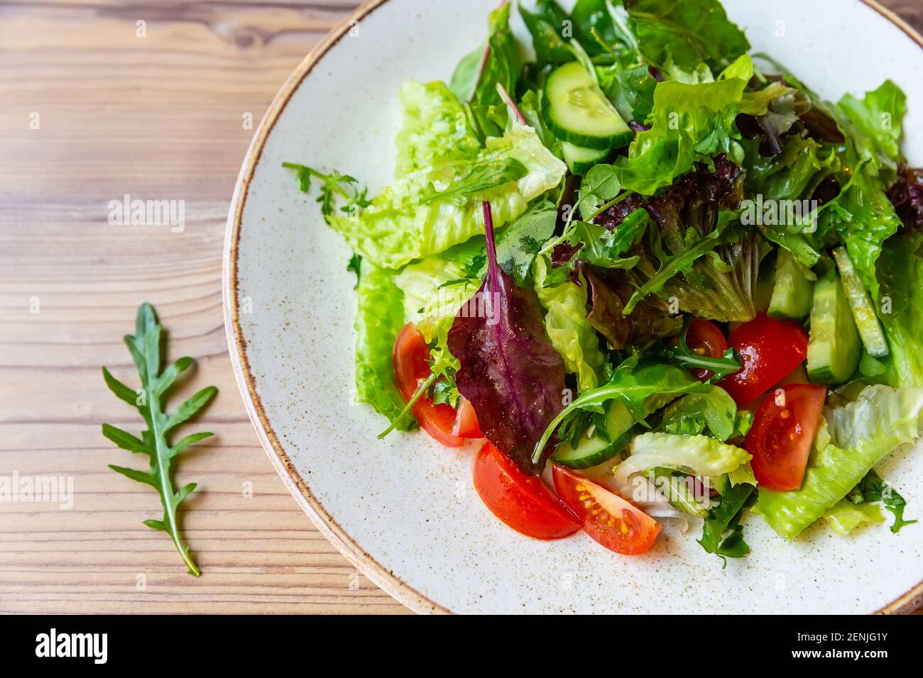 Frischer Gemüsesalat mit Tomaten, Gurken, Zwiebeln, Spinat, Salat und Rucola auf weißem Teller. Schüssel Salat mit Gemüse auf Holztisch. Stockfoto