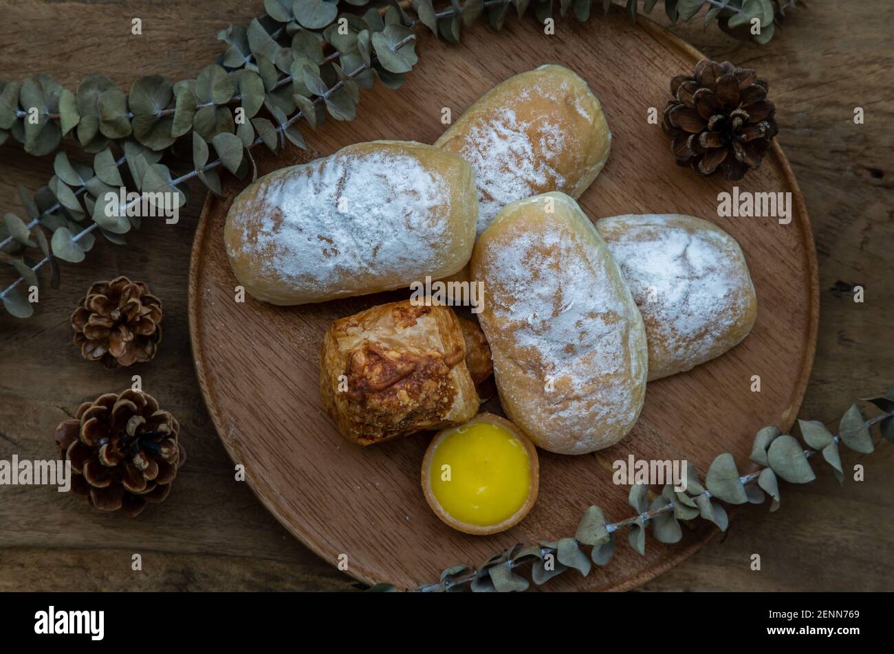 Hausgemachte süße Sahne-Softbrötchen, gebackene Croissant-Wurst und Zitronentarte mit getrockneten Blumen auf runden Holzteller auf rustikalem Holzboden. Bäckerei und d Stockfoto