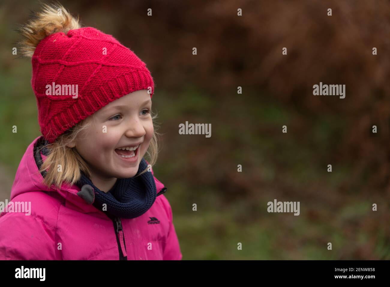 Junges Mädchen in rosa Wintermantel & roter Hut Wandern im Wald, lachend, grüner Hintergrund, Winterspaziergang im Wald, lachendes Mädchen im Wald, Hoffnung Stockfoto