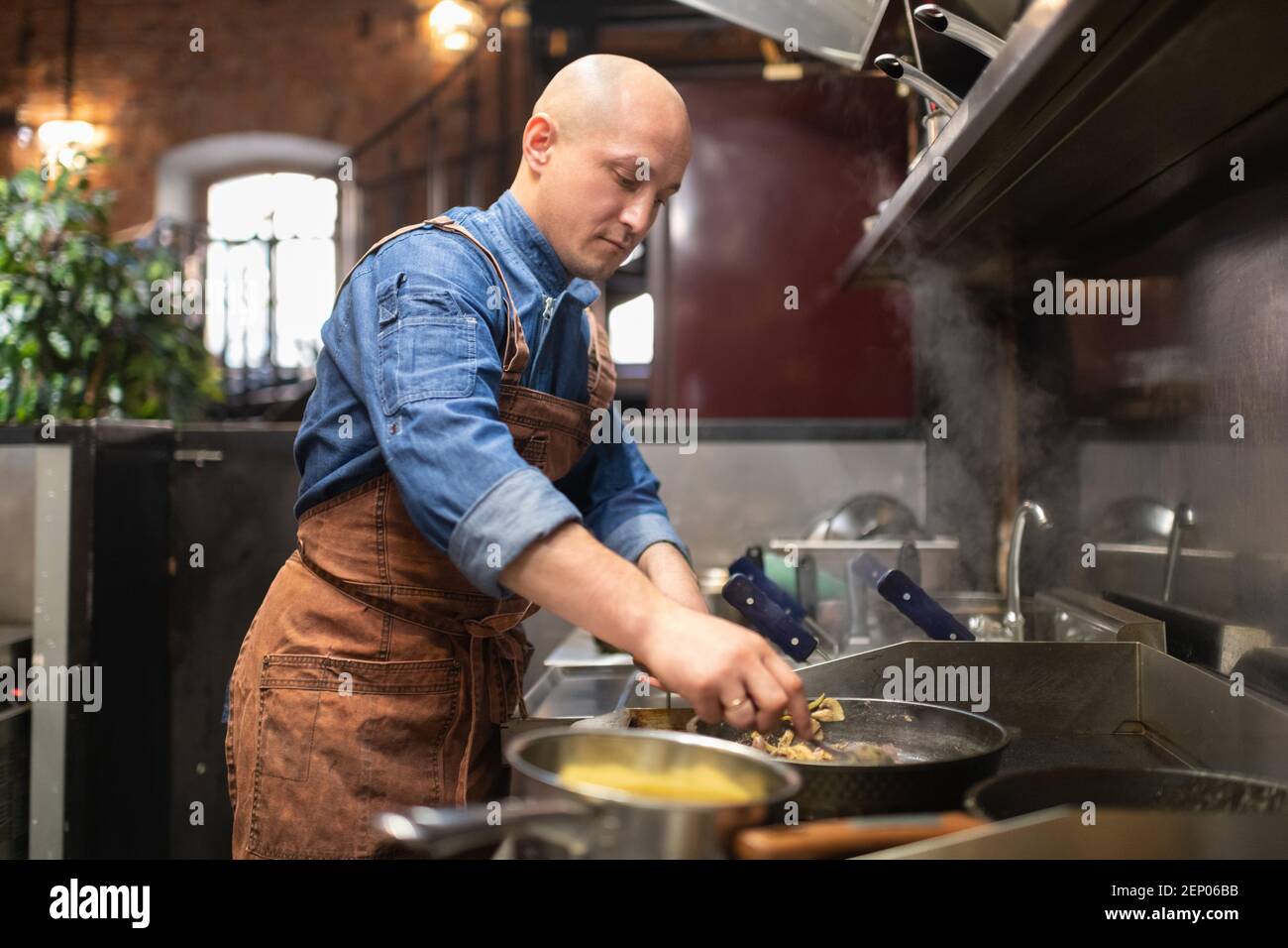 Glatze Koch in Schürze Rühren Gericht auf Bratpfanne, während Kochen von Essen in der Café-Küche Stockfoto