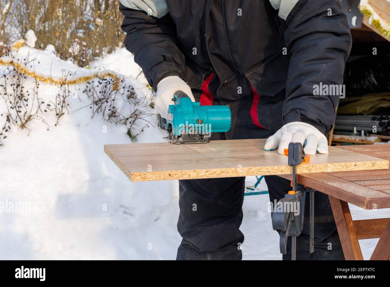 Ein männlicher Arbeiter schneidet eine Holzplatte, die mit Klammern an einer Werkbank befestigt ist, mit einer kreisförmigen Scheibe auf einer Batterie. Stockfoto