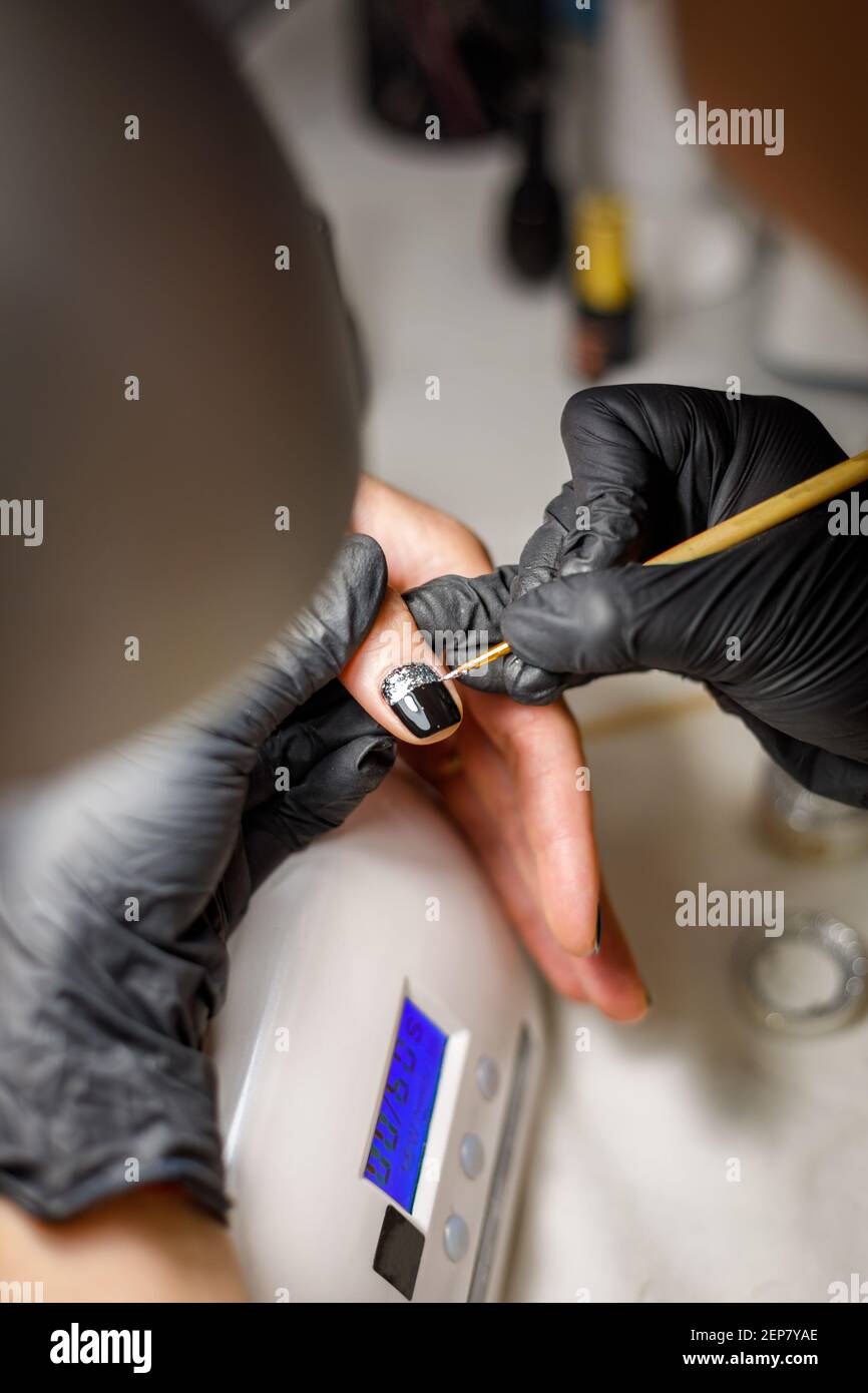 In einem Schönheitssalon macht der Meister eine Maniküre, trägt ein Muster auf den Nagel mit einem Pinsel, schwarzer Lack mit einem goldenen Muster. Stockfoto