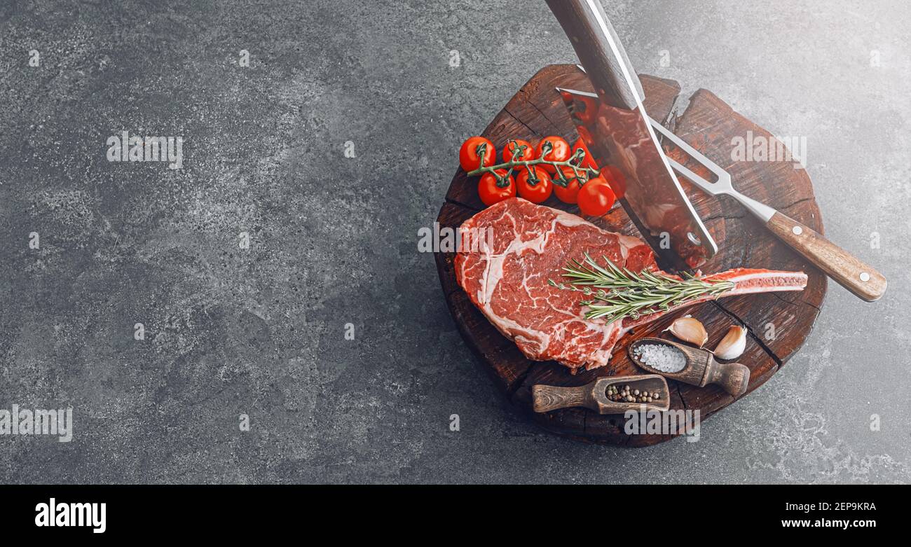 Auf einem Holzblock für Fleisch ein frisches rohes tamahawk Steak oder ein Cowboy Steak mit einer Metzgerhackaxt für Fleisch, daneben eine Mischung aus Paprika und Stockfoto