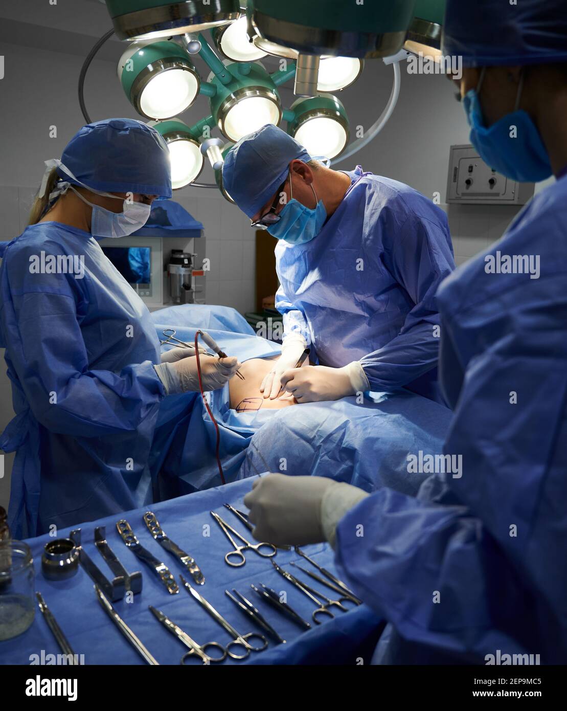 Operationsteam in sterilen Handschuhen und Schutzmasken tun ästhetische Chirurgie im Operationssaal. Assistent steht neben OP-Tisch mit medizinischen Werkzeugen. Konzept der Medizin und plastische Chirurgie. Stockfoto