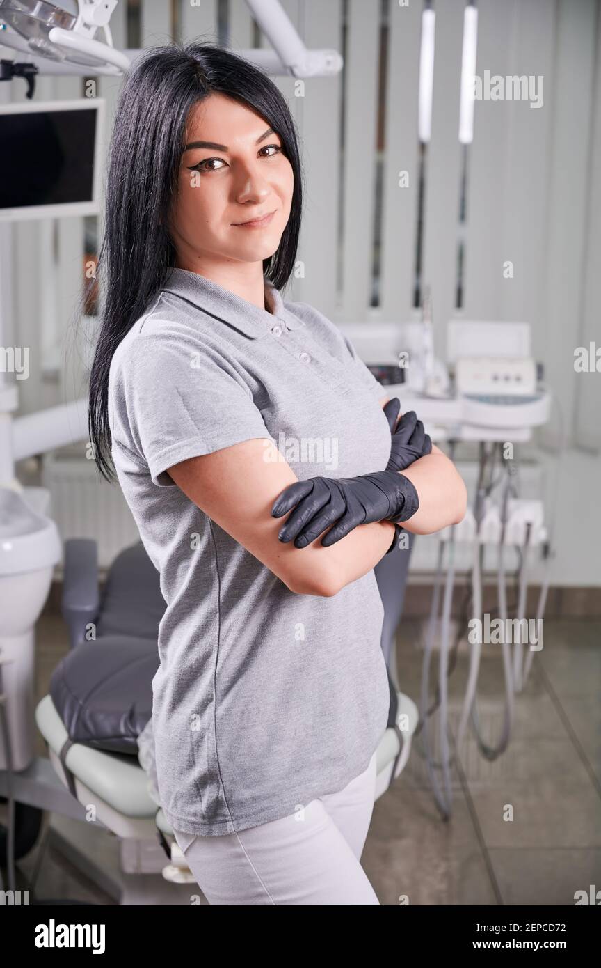 Porträt einer Zahnärztin, die in einem gut ausgestatteten Büro steht, lächelt, Uniform und schwarze Handschuhe trägt. Konzept der Gesundheitsversorgung und kieferorthopädische Dienstleistungen Stockfoto