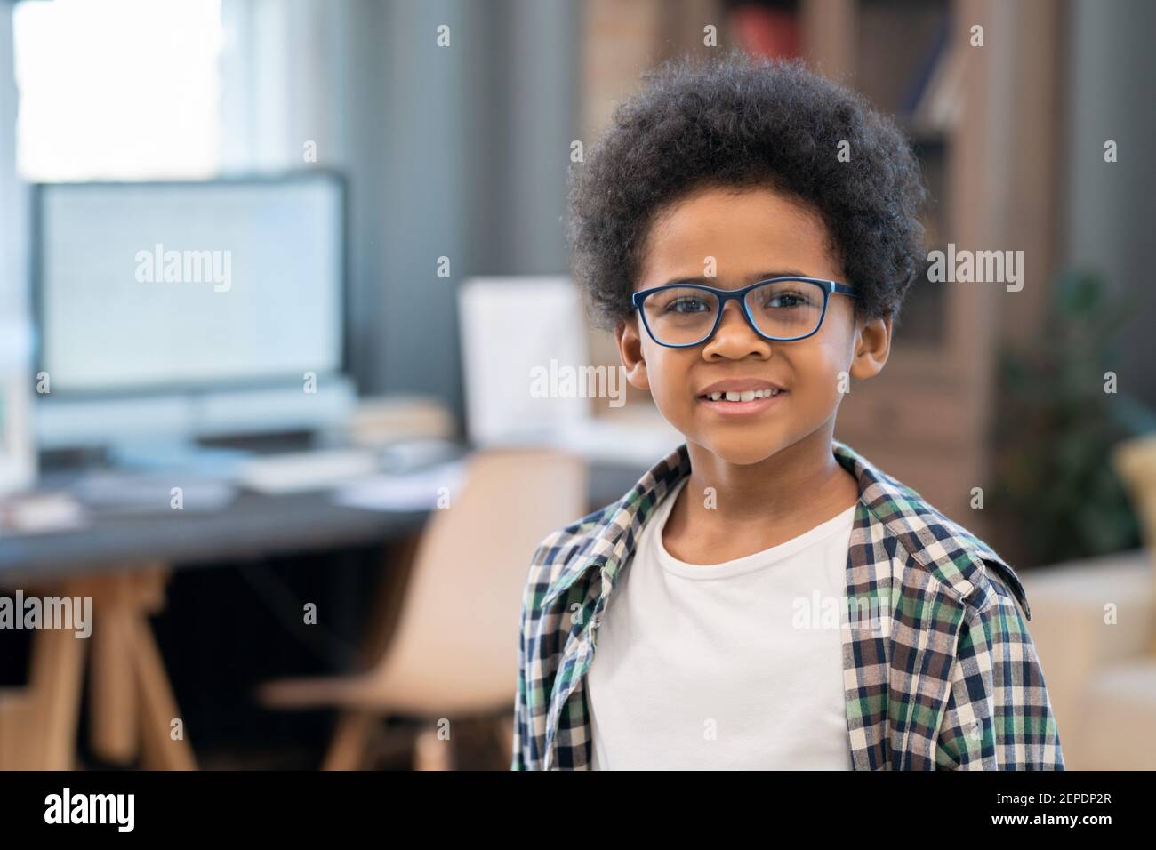 Liebenswert Mixed-Race Schuljunge in Casualwear und Brillen stehen vor Von Kamera gegen Tisch mit Computer und Blick auf Sie Stockfoto