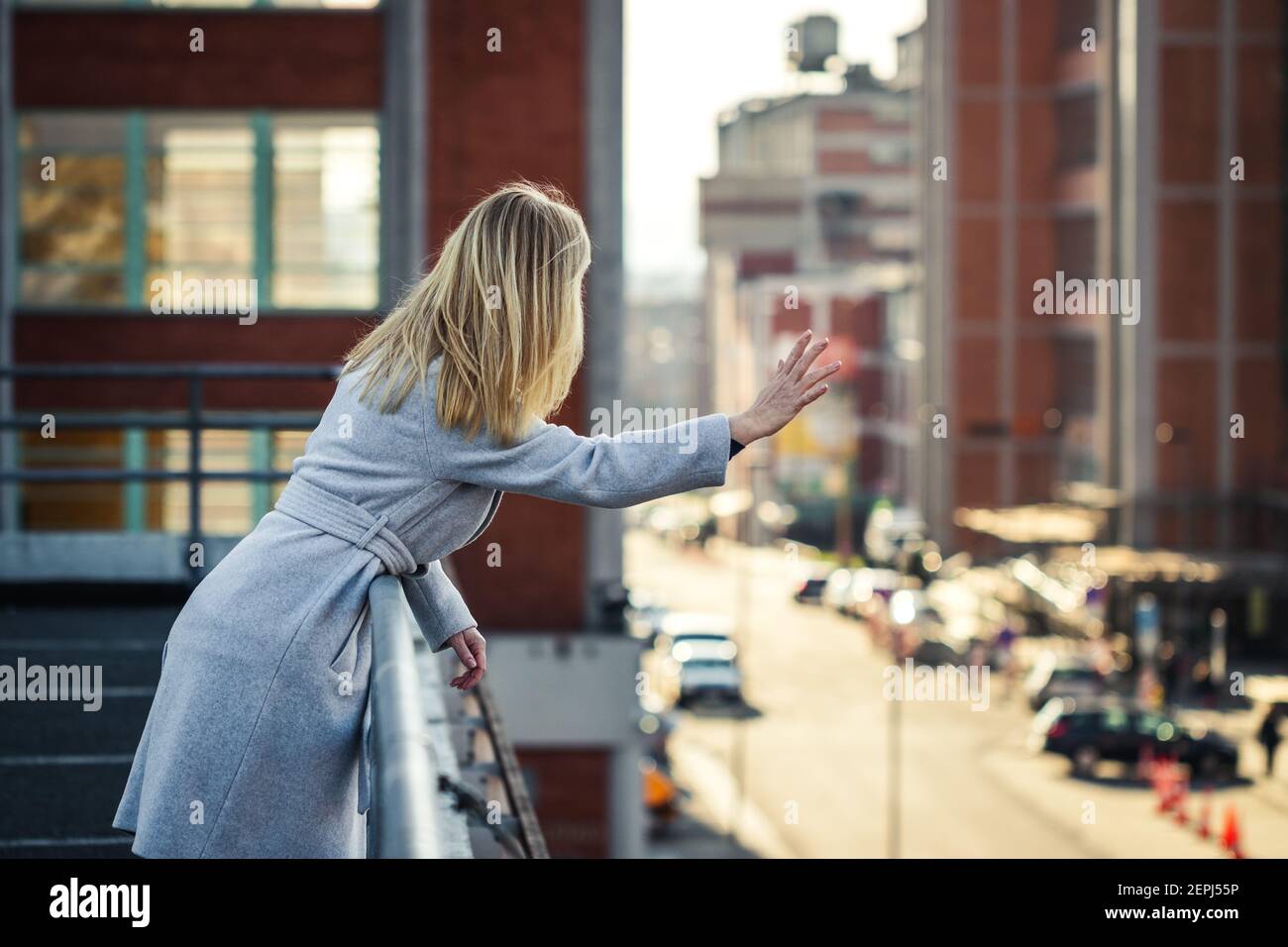 Frau winkte von der Terrasse zu den Leuten auf der Straße. Stadtbild Hintergrund. Blonde Haare Frau trägt stilvolle grauen Mantel in der Stadt Stockfoto