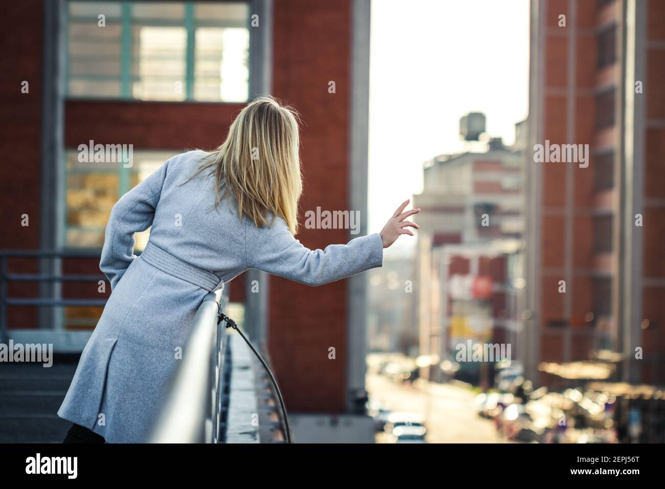 Modische Frau winkt von der Terrasse zu den Menschen in der Stadt Straße. Stadtbild Hintergrund. Blonde Haare Frau trägt stilvolle grauen Mantel in der Stadt Stockfoto