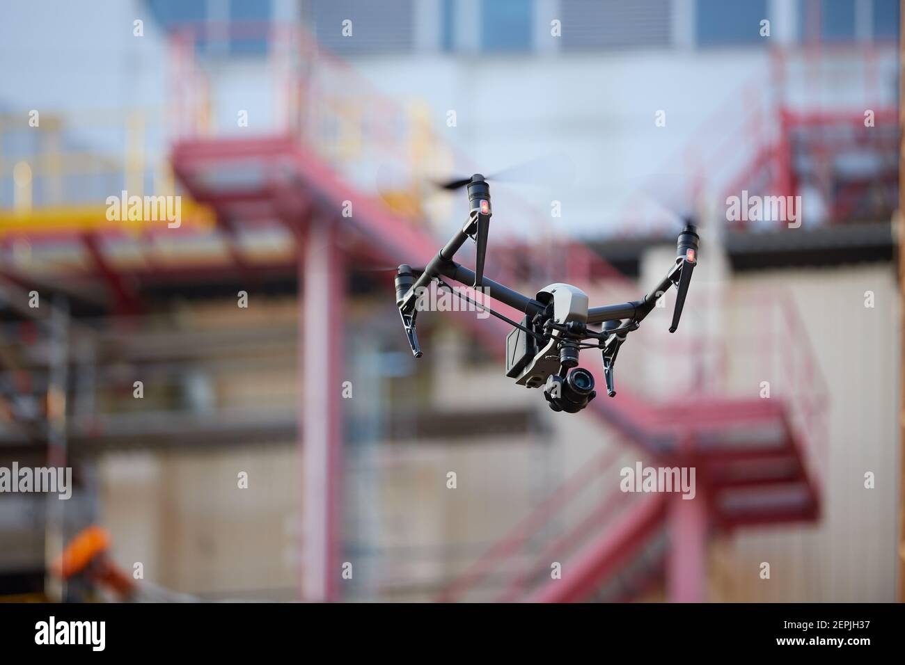 Schwebender schwarzer Quadrocopter mit leistungsstarker Kamera gegen unscharfe iindustrielle Bauweise. Sichtprüfung in der Industrie. Stockfoto