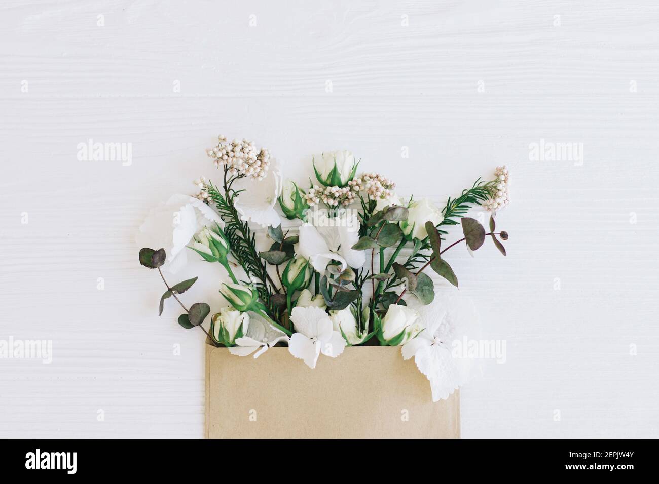 Stilvolle Rosen, Hortensien und Eukalyptuszweige wachsen aus Handwerk Umschlag auf weißem Holz, flach legen mit Kopie Raum. Blumen Grußkarte und Blume Stockfoto