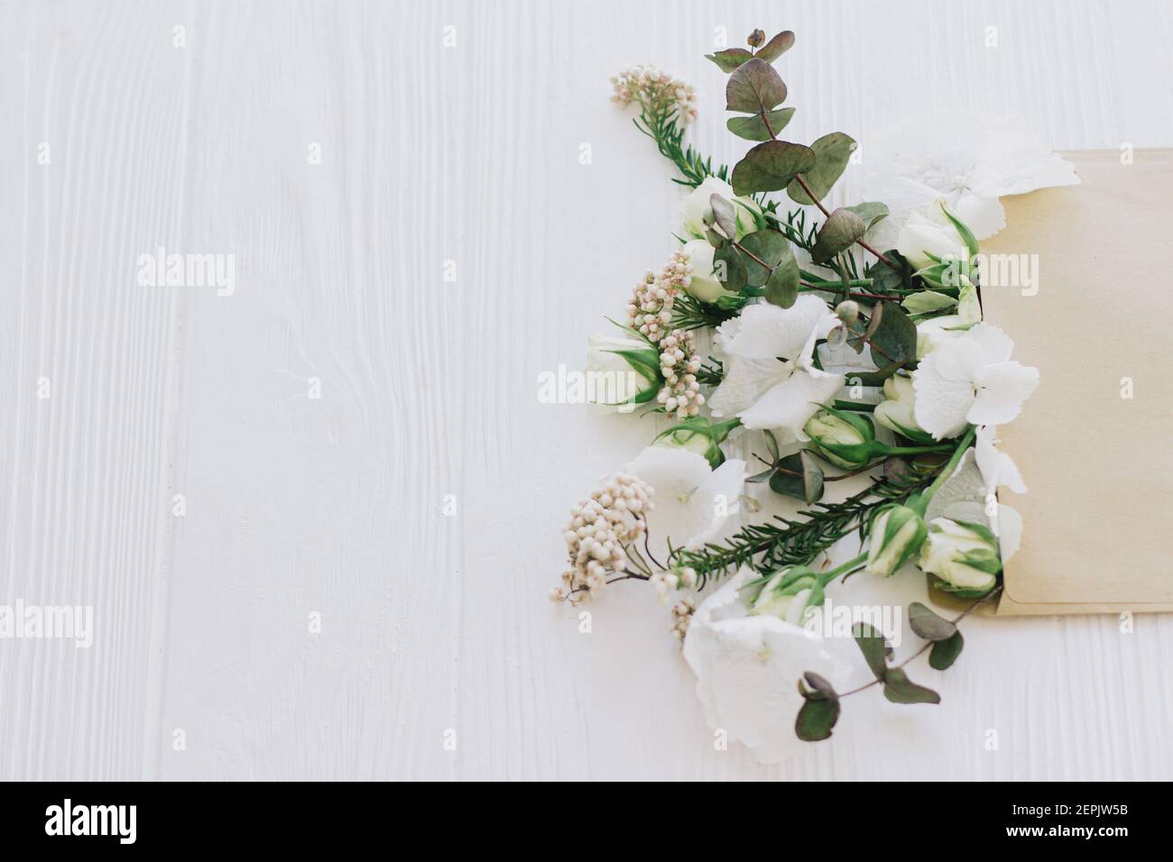 Stilvolle Rosen, Hortensien und Eukalyptuszweige wachsen aus handgefertigte Umschlag auf weißem Holz mit Kopieplatz. Kreatives Frühlingsbild, florale Begrüßung ca. Stockfoto