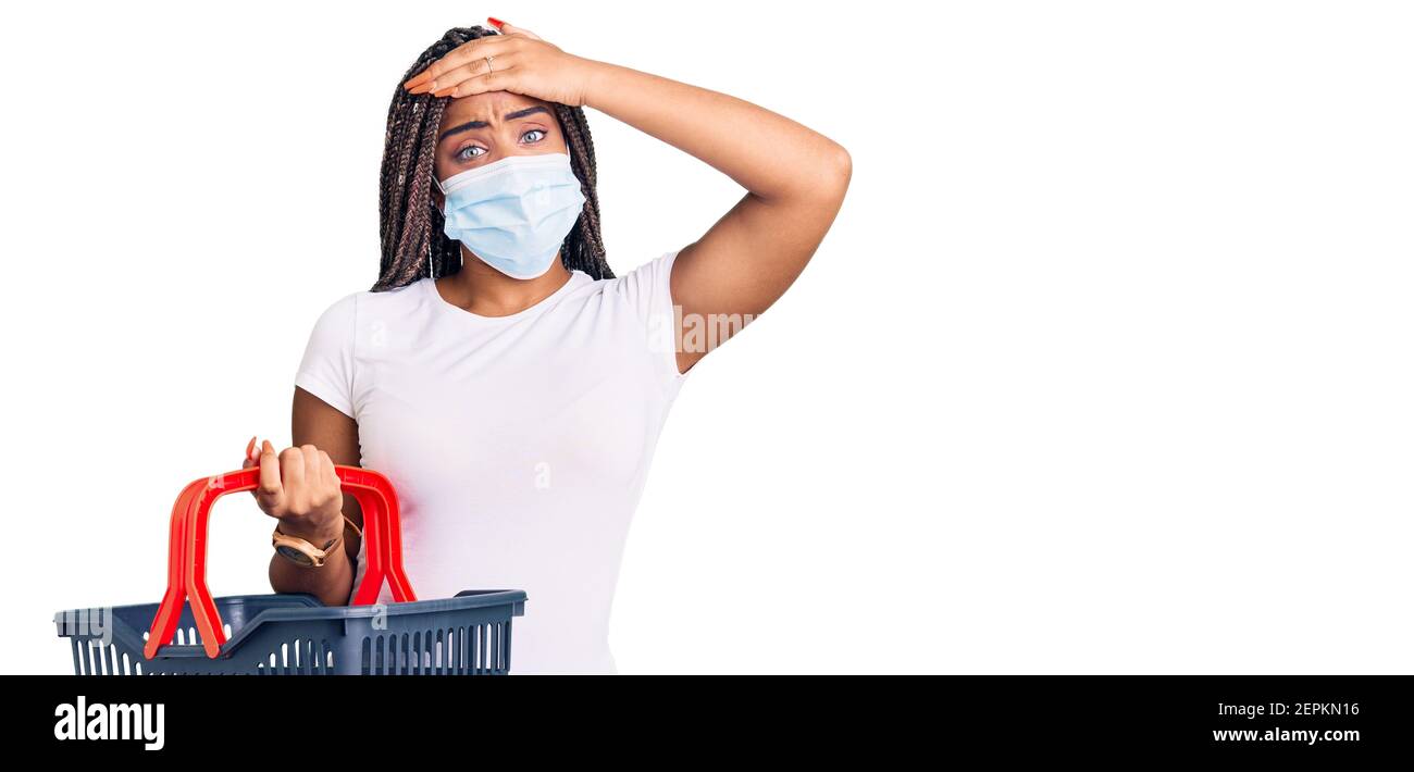Junge afroamerikanische Frau mit Zöpfen tragen Warenkorb und medizinische Maske gestresst und frustriert mit Hand auf Kopf, überrascht und wütend Gesicht Stockfoto