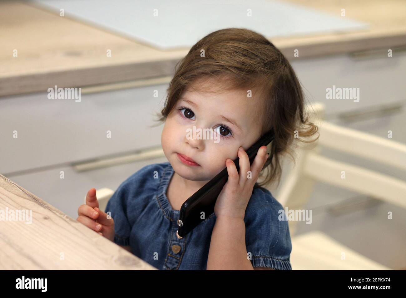 Nettes kleines dunkelhaariges Mädchen 1,5-2,5 in einem Jeanskleid spricht  mit jemandem auf einem Smartphone in der Küche. Baby vermissen  Kommunikation mit Verwandten. Se Stockfotografie - Alamy
