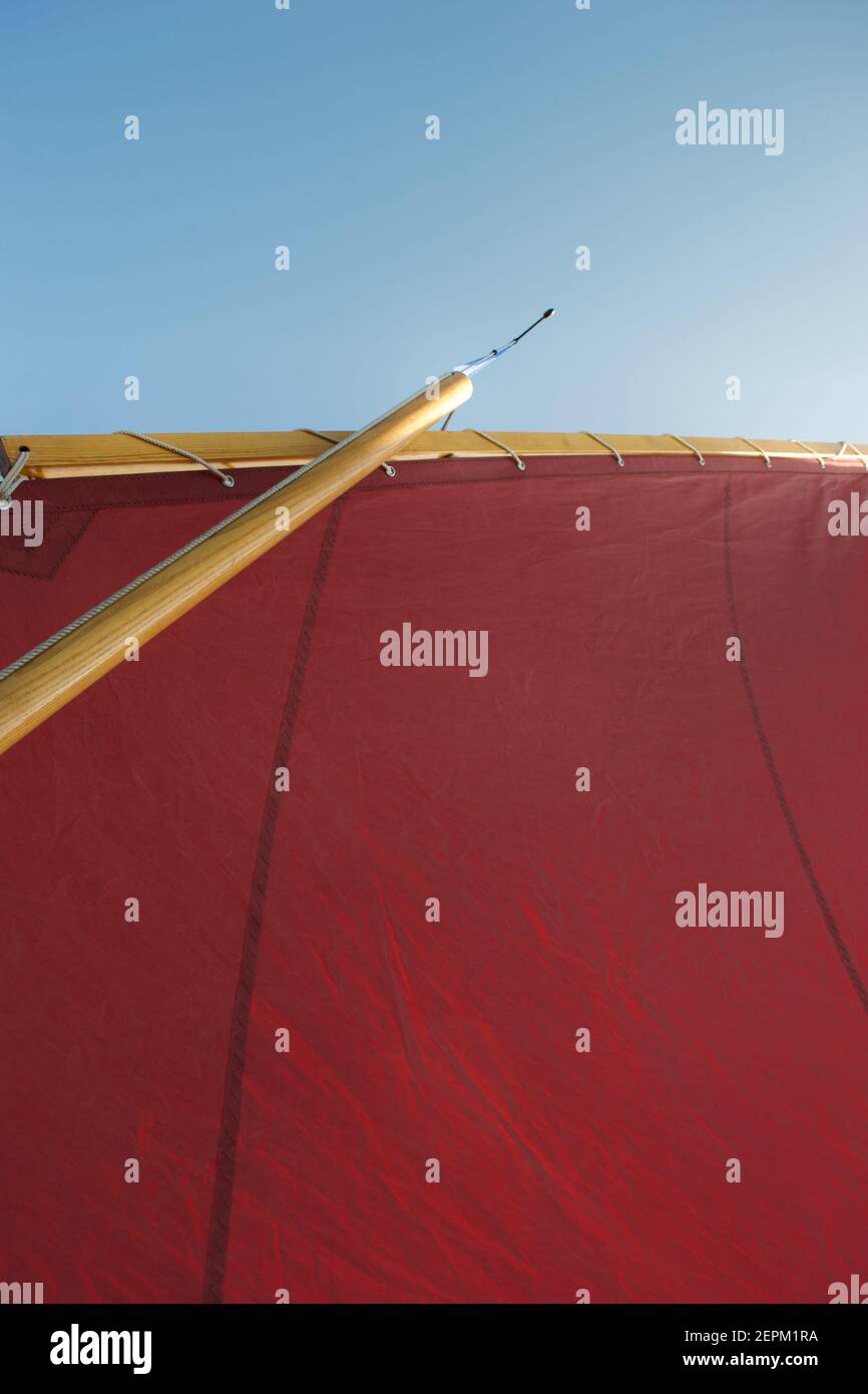 Blick auf die Takelage eines gaffig manipulierten Segeljinghy: Rotes Lugsail-Segel, Holzmast, Burgee und strahlend blauer Himmel über Stockfoto