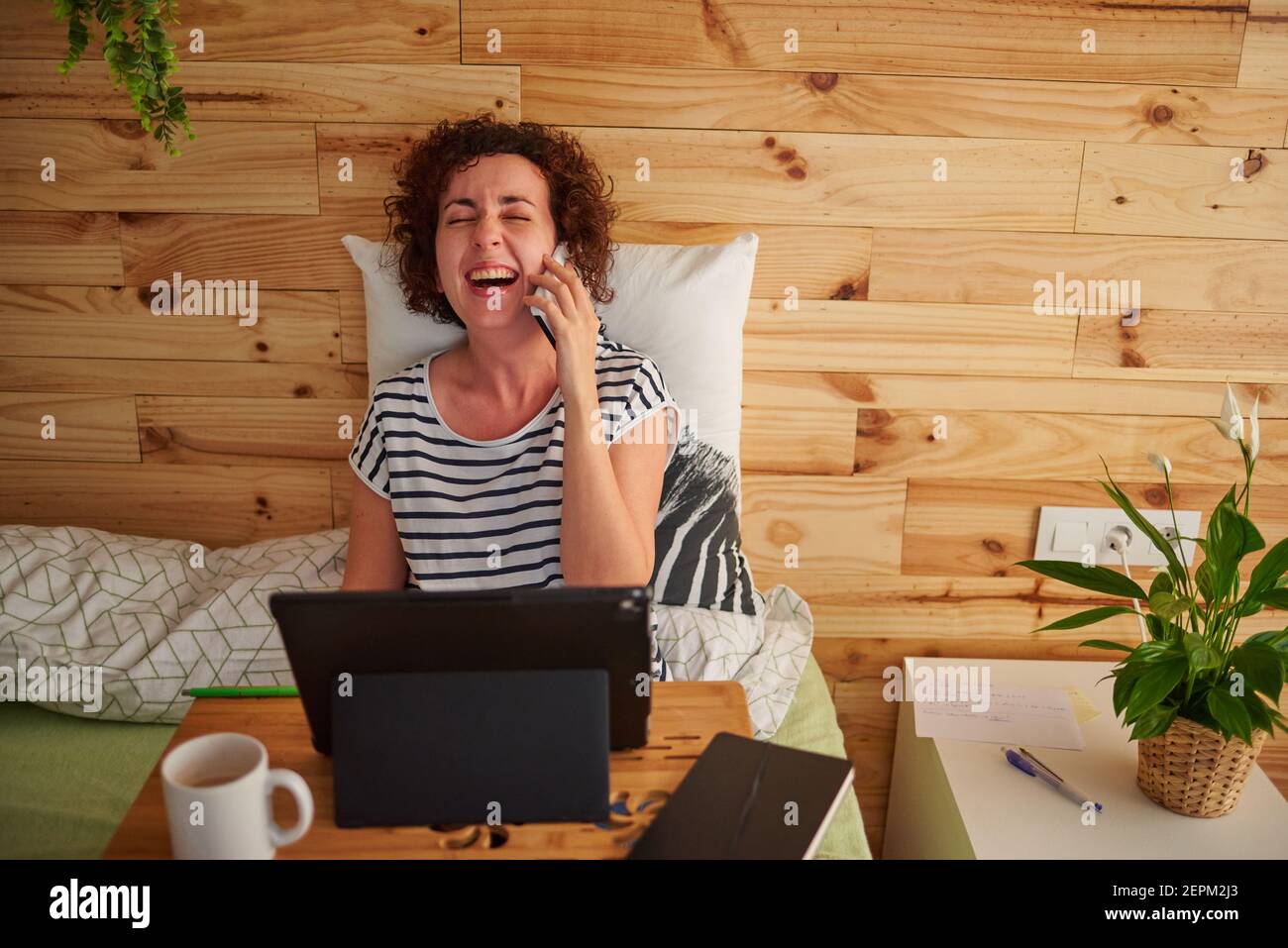 Eine lockige kaukasische Frau lacht, während sie während einer Arbeitsschicht von zu Hause aus am Telefon spricht. Sie sitzt auf dem Bett mit einem Klapptisch, dass sh Stockfoto