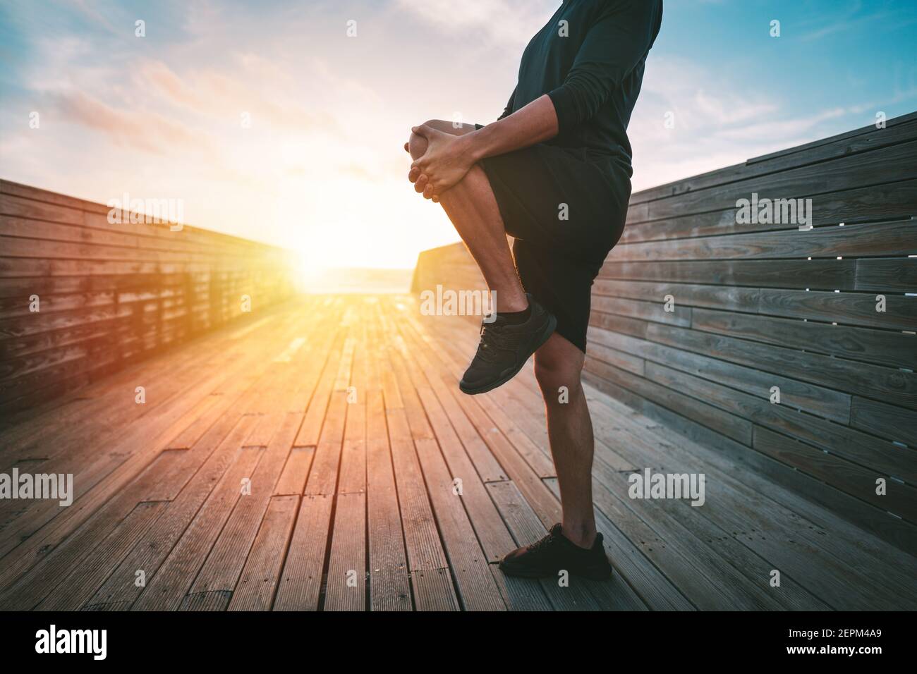 Gesunder junger Mann, der sich vor dem Training im Freien bei Sonnenuntergang oder Sonnenaufgang die Beine streckt. Dehnung des Musculus gluteus maximus. Läufer bereitet sich auf den Lauf vor. Stockfoto