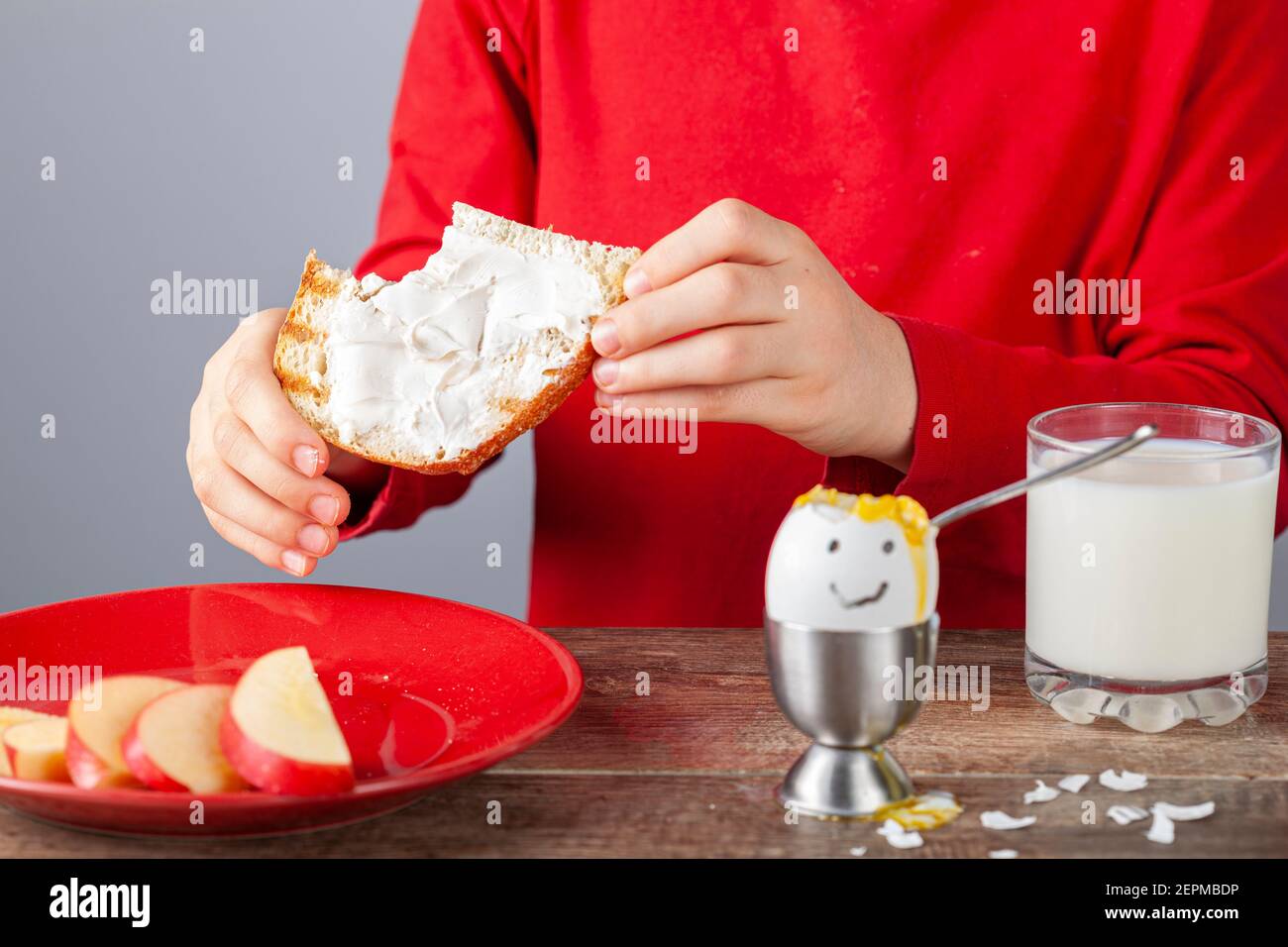 Frühstückstisch mit einfachen ausgewogenen nahrhaften Zutaten. Ein weich gekochtes Ei mit bemaltem Gesicht, eine Scheibe geröstetes Brot mit Frischkäse-Aufstrich Stockfoto