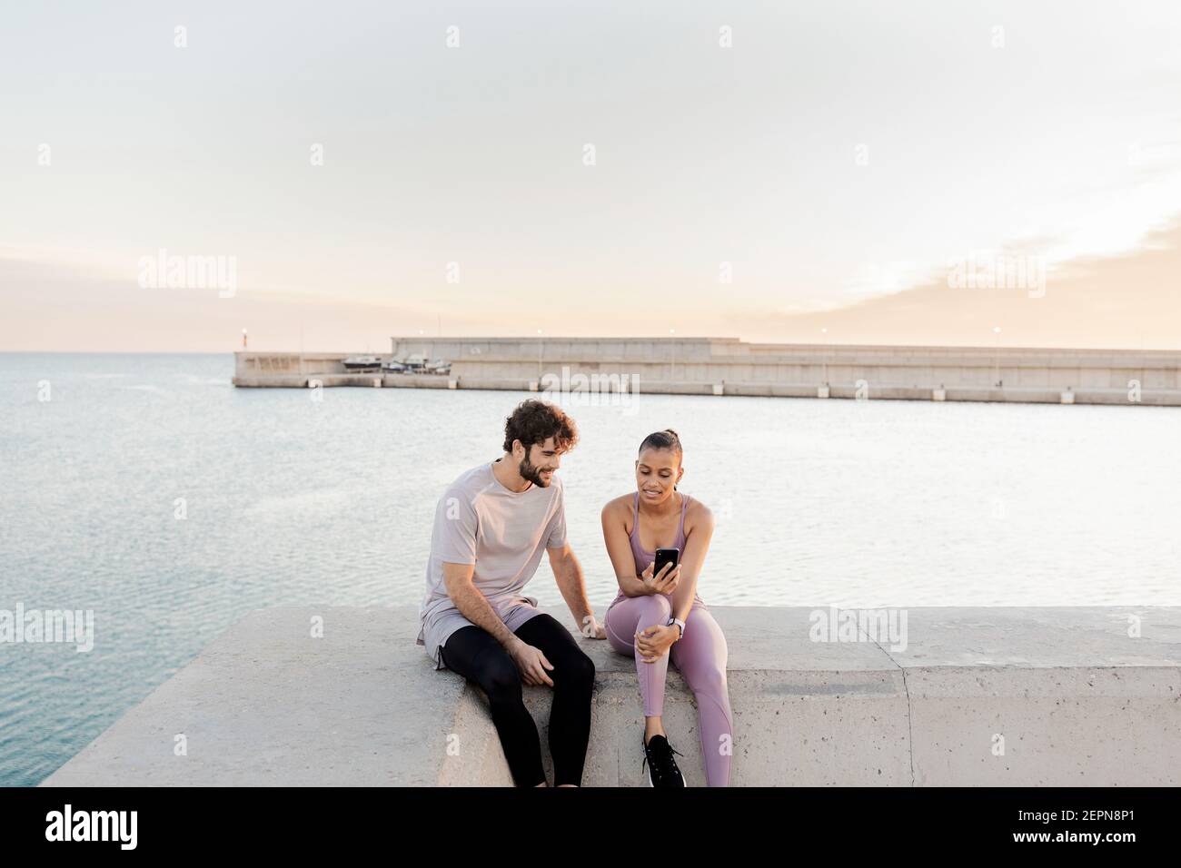 Junge froh Sportler mit ethnischen weiblichen Partner in Sportkleidung Während die Zeit zusammen mit dem Handy gegen Meer nach Schulung Stockfoto