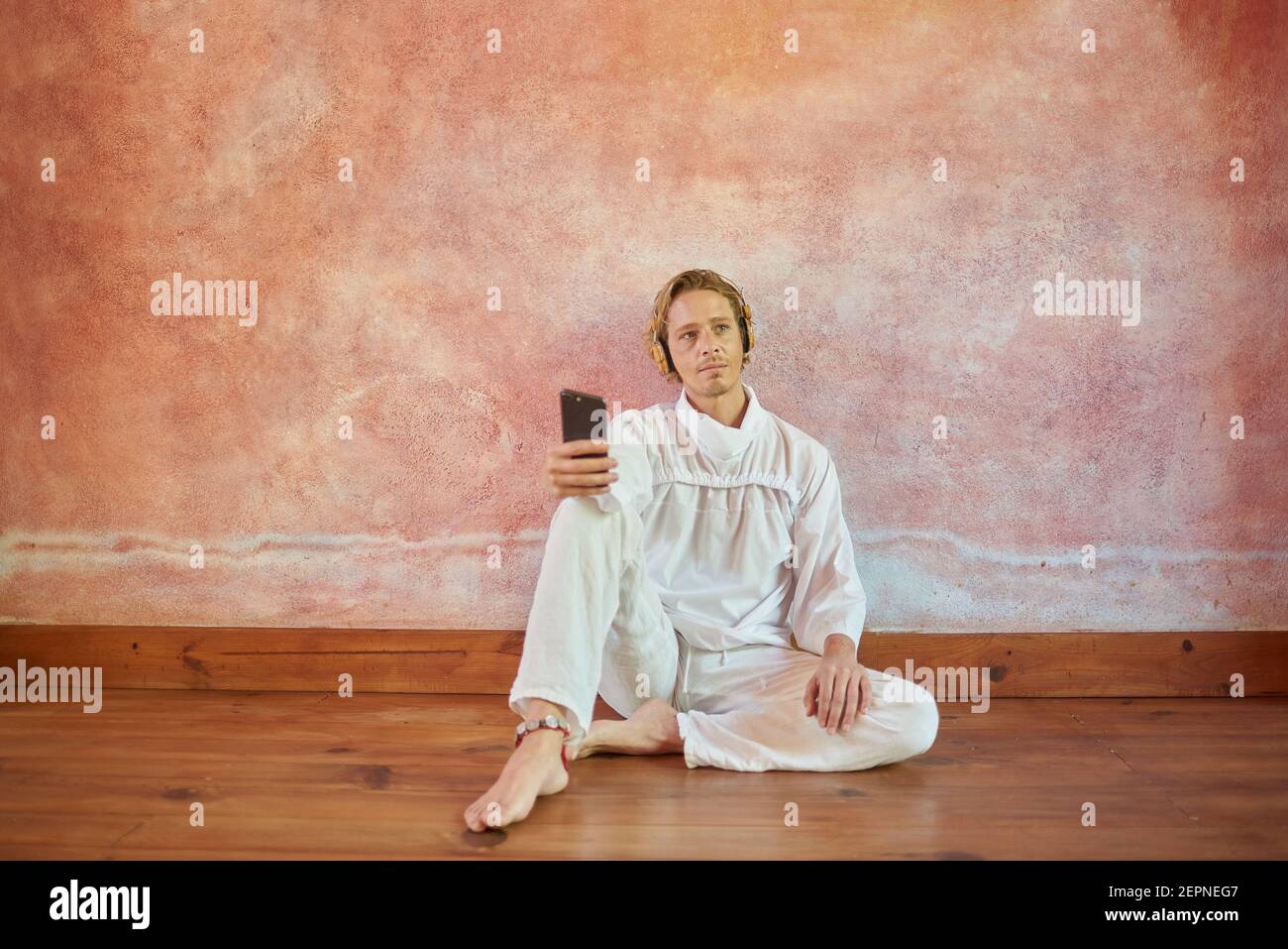 Junge achtsame Mann in weißem Outfit mit Handy sitzend lehnend An einer Wand hören Yoga-Kurs von Headset suchen Weg Stockfoto