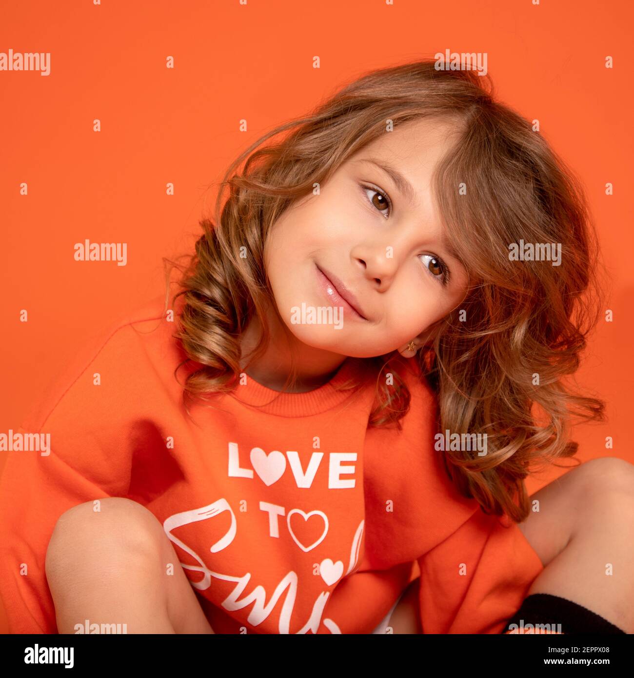 Nahaufnahme Foto von einem netten schönen Baby Mädchen 6-7 Jahre alt, umarmte ihre Knie auf orangefarbenem Hintergrund. Wegschauen Stockfoto