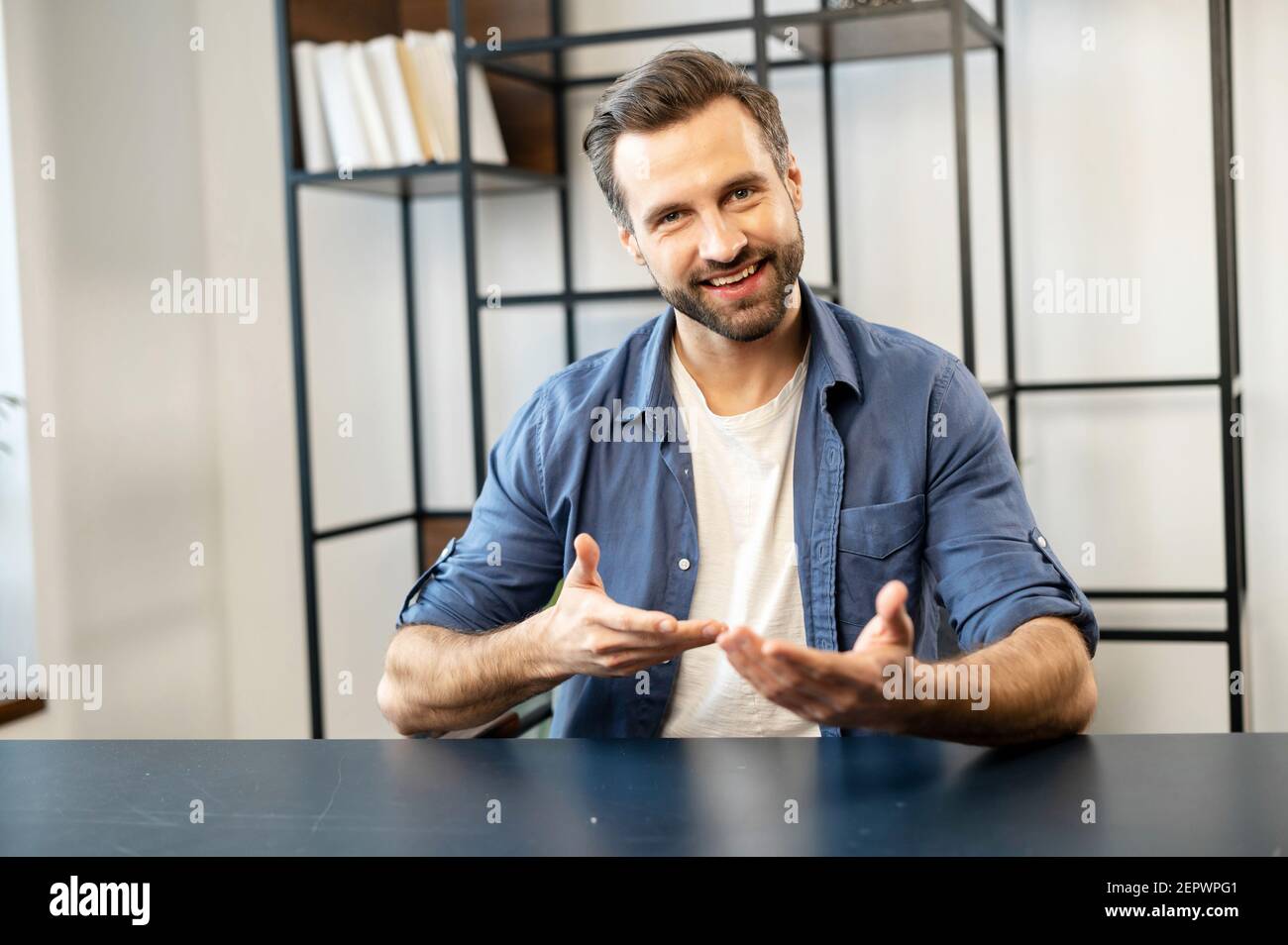 Junger bärtiger Hipster in lässiger Kleidung sitzt am Schreibtisch, Blick auf die Kamera, Hände bewegen, jemandem sagen, mehr über sich selbst erzählen oder präsentieren die Idee, während auf Business-Video-Anruf Stockfoto