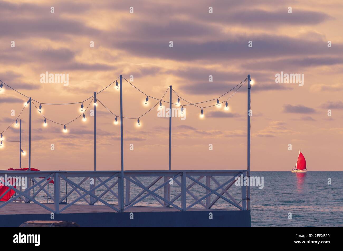 Romantischer Pier bei Sonnenuntergang mit glühenden Glühbirnen, Seitenblick. Eine Yacht mit rotem Segel steht am Horizont. Stockfoto