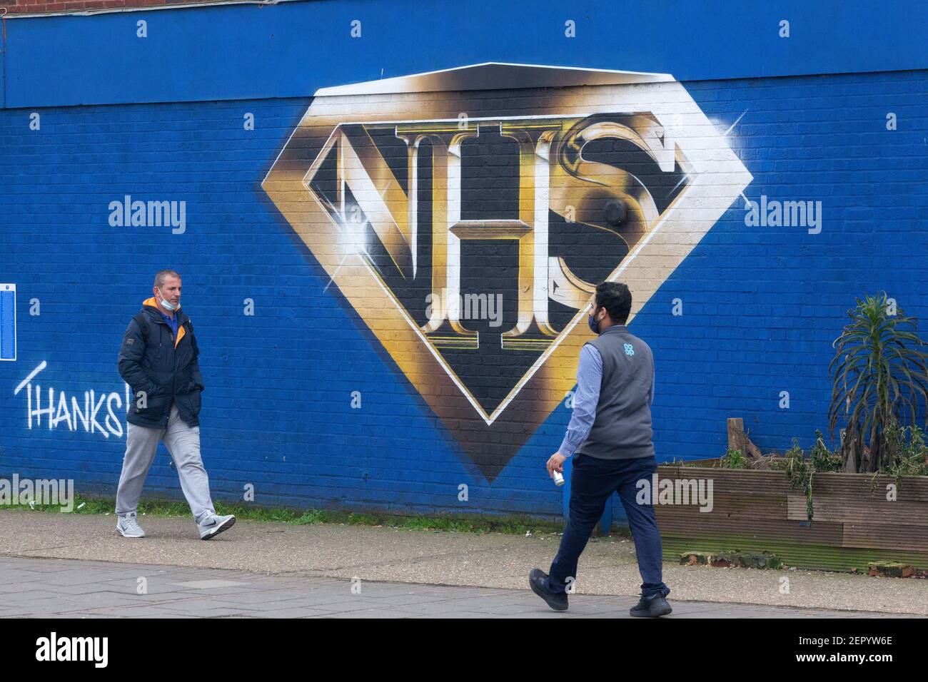 London, Großbritannien, 28. Februar 2021: Ein Wandgemälde in Tulse Hill zeigt das NHS-Logo im Stil eines Superhelden-Emblems. Passanten nehmen Übung oder Schlange für den Co-op-Laden um die Ecke, einige tragen Gesichtsmasken und alle sozial distanziert. Anna Watson/Alamy Live News Stockfoto