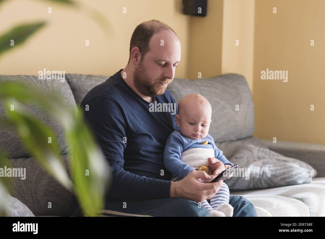 Kaukasischer Vater im Gespräch auf Handy, um online mit seinem neugeborenen Baby zu arbeiten. Alleinerziehende Vater kümmert sich um seinen Sohn. Glückliche Familie und Arbeit zu Hause Stockfoto