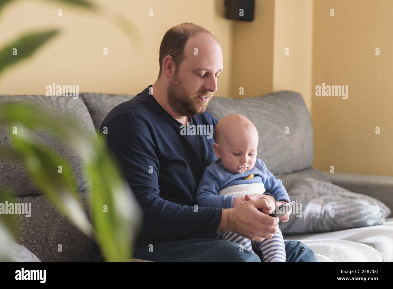 Kaukasischer Vater im Gespräch auf Handy, um online mit seinem neugeborenen Baby zu arbeiten. Alleinerziehende Vater kümmert sich um seinen Sohn. Glückliche Familie und Arbeit zu Hause Stockfoto