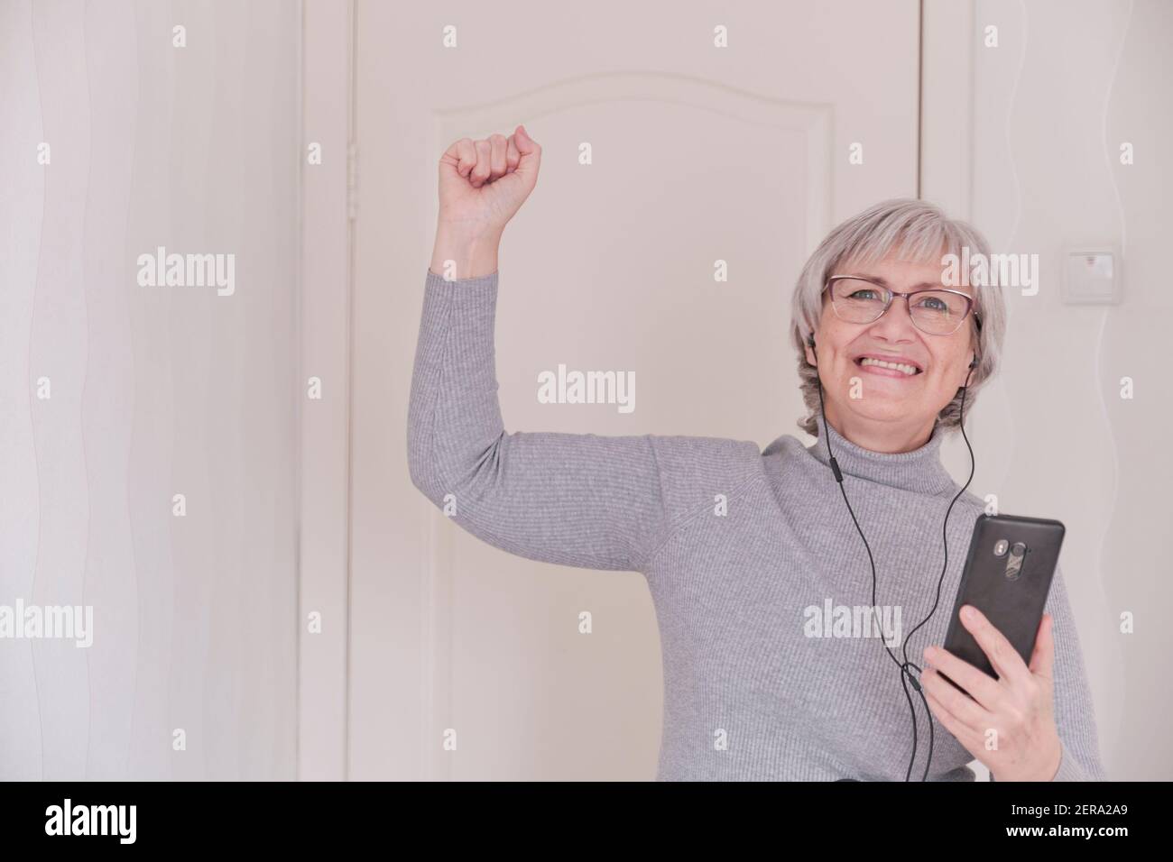 Eine grauhaarige ältere Frau mit Brille, in Kopfhörern, in einem grauen Rollkragen, die zu Hause auf dem Handy telefoniert und enttäuscht ist. Menschen verbinden sich Stockfoto