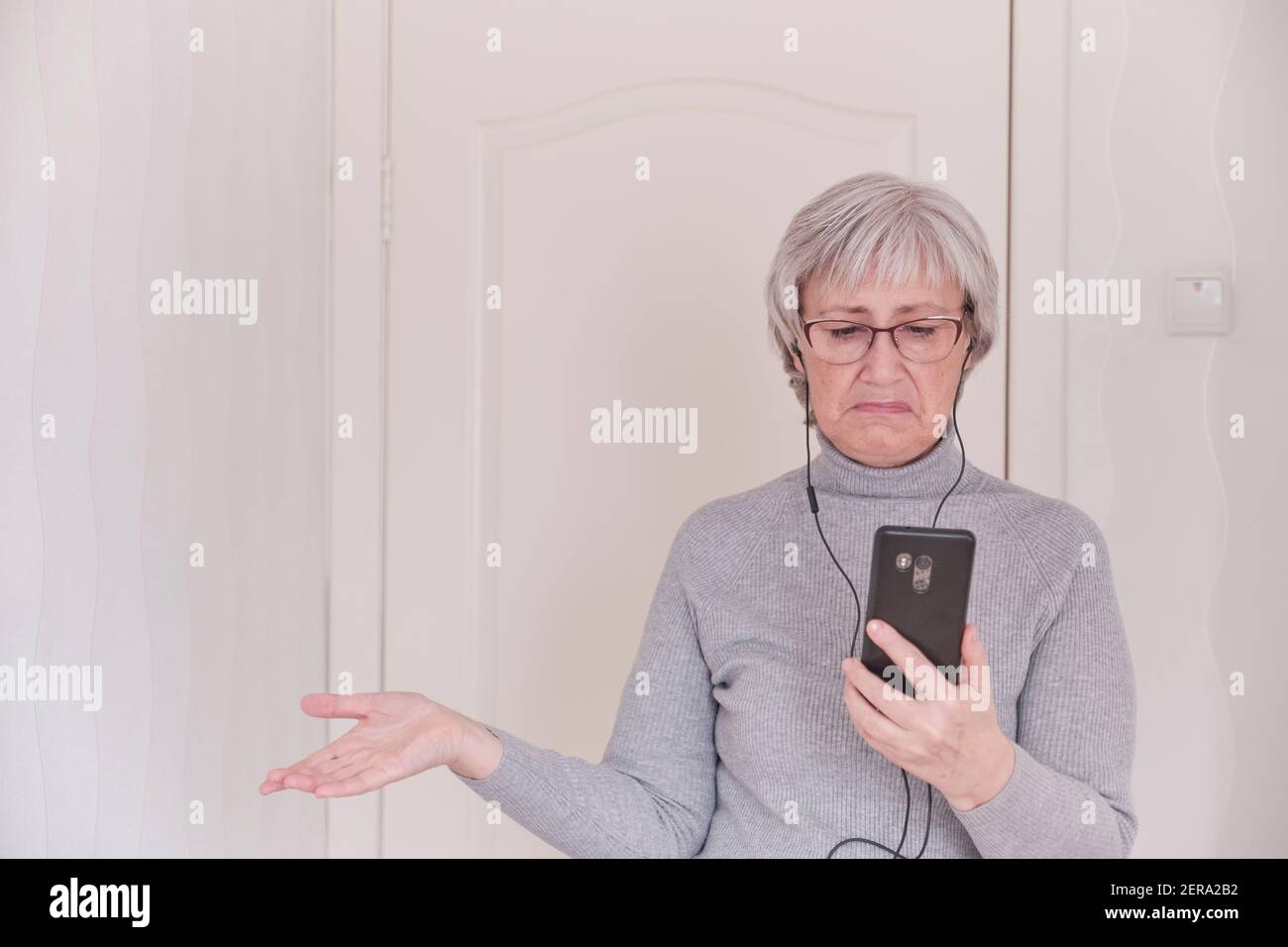 Eine grauhaarige ältere Frau mit Brille, in Kopfhörern, in einem grauen Rollkragen, die zu Hause auf dem Handy telefoniert und enttäuscht ist. Menschen verbinden sich Stockfoto