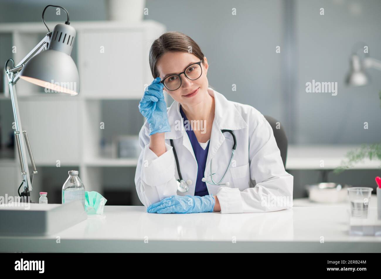 Portrait einer niedlichen Ärztin in blauen medizinischen Latexhandschuhen am Tisch, für die Aufnahme von Patienten. Ein vertrauenswürdiges Gesicht und ein gepflegtes Aussehen Stockfoto