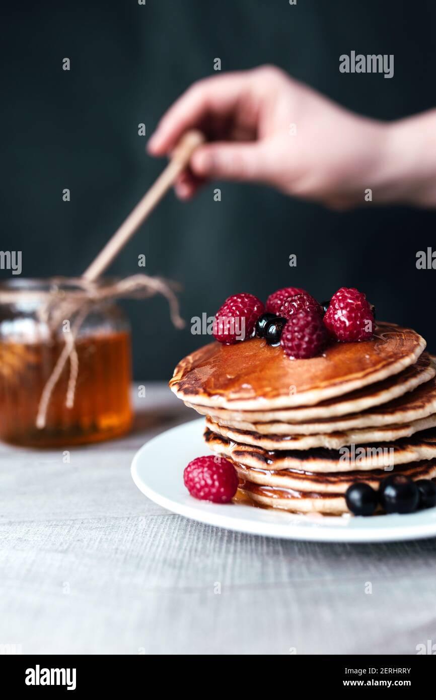 Pfannkuchen mit Beeren und Honig auf weißem Teller, Hand halten Löffel im Glas, Holztisch. Hochwertige Fotos Stockfoto
