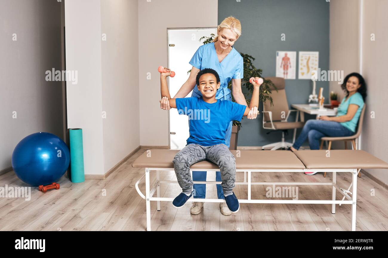 Afroamerikanischer Junge trainiert mit Frau Physiotherapeutin mit Kurzhanteln im Reha-Zentrum. Lächelndes Kind mit den Händen nach oben, Hanteln halten Stockfoto