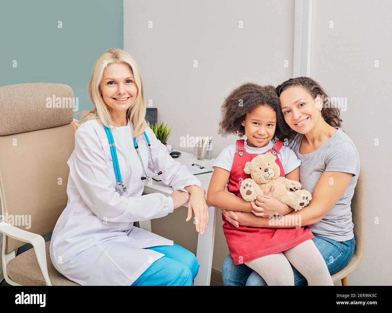 Afroamerikanisches Mädchen mit ihrer Mutter und Kinderärztin sitzen in einer Arztpraxis, lächeln und schauen in die Kamera. Rücksprache mit chil Stockfoto